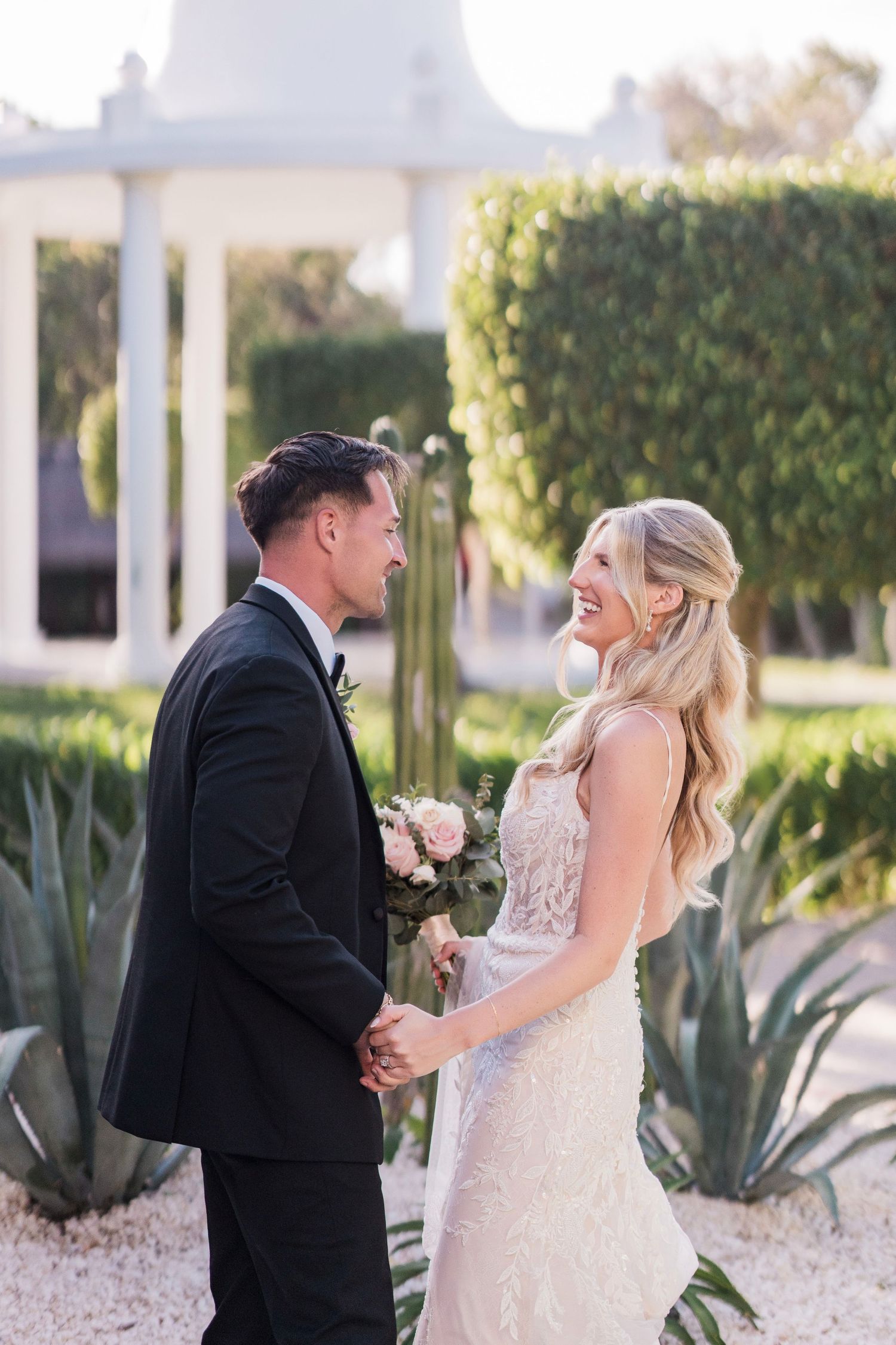 A smiling bride in a lace gown holds a pink bouquet while holding hands with her groom in a black tuxedo outdoors.