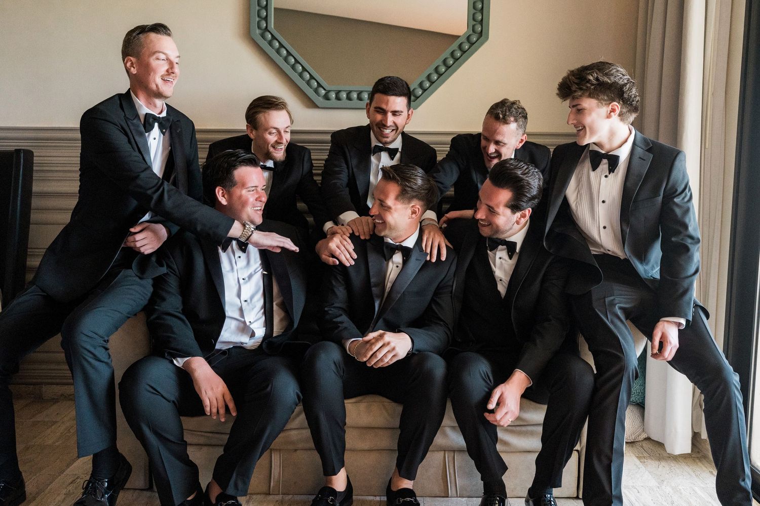 Group of eight groomsmen in black tuxedos laughing together while seated in an elegant hotel room.