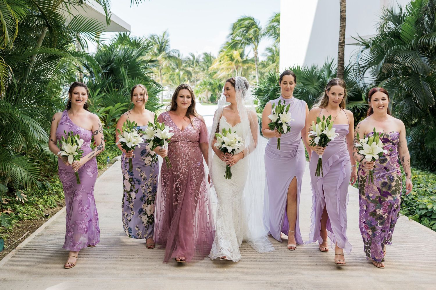 Bride and bridesmaids in purple dresses holding white floral bouquets walking on a tropical resort pathway.