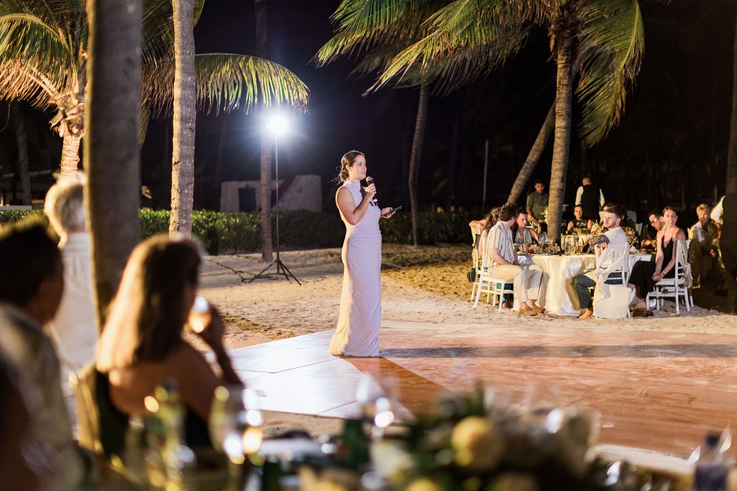 Bride in white gown giving speech at outdoor beach wedding reception at night with palm trees and guests seated at tables.