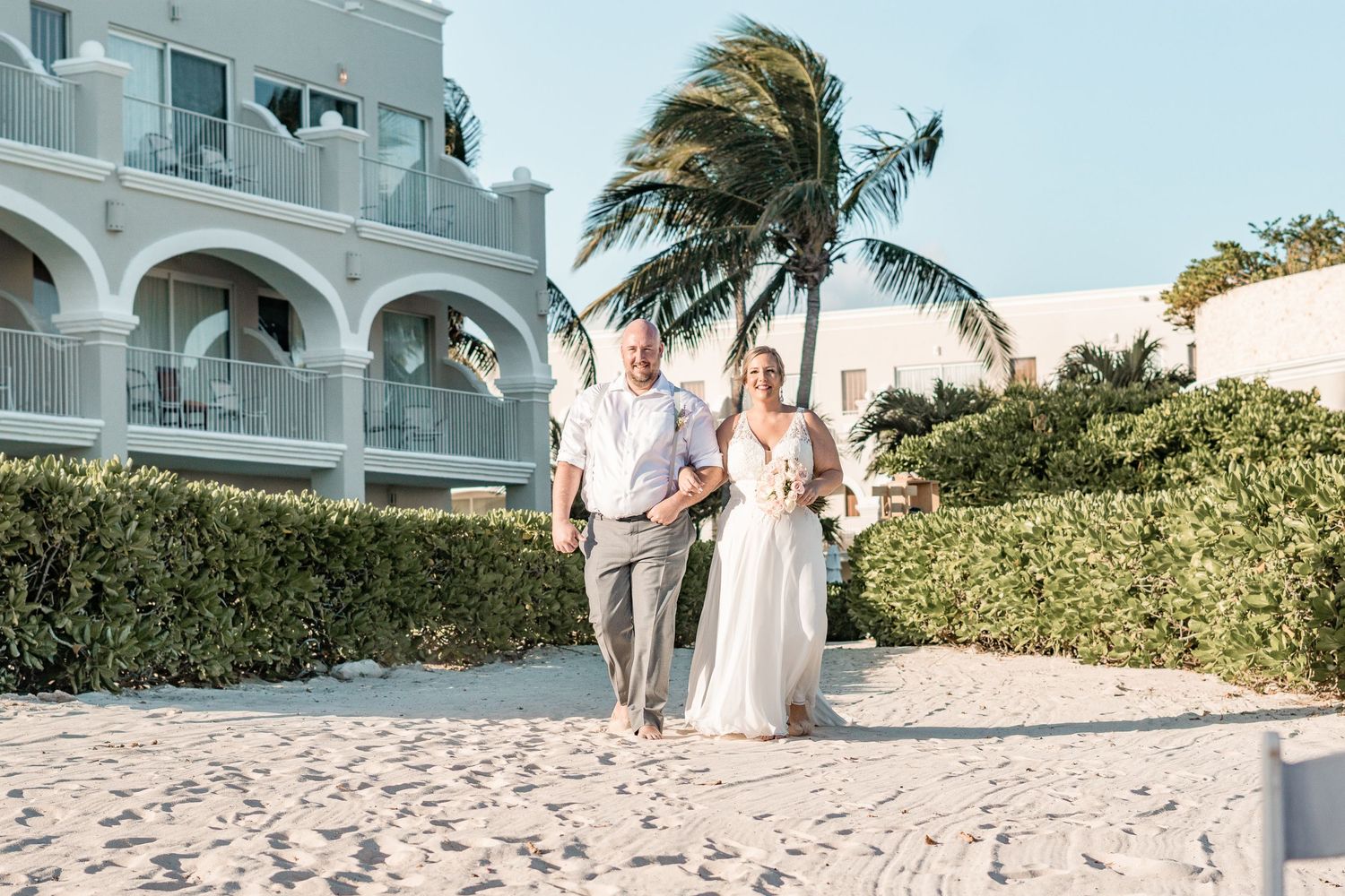 Bride and groom walk hand-in-hand on white sand beach with tropical palm trees and resort backdrop.