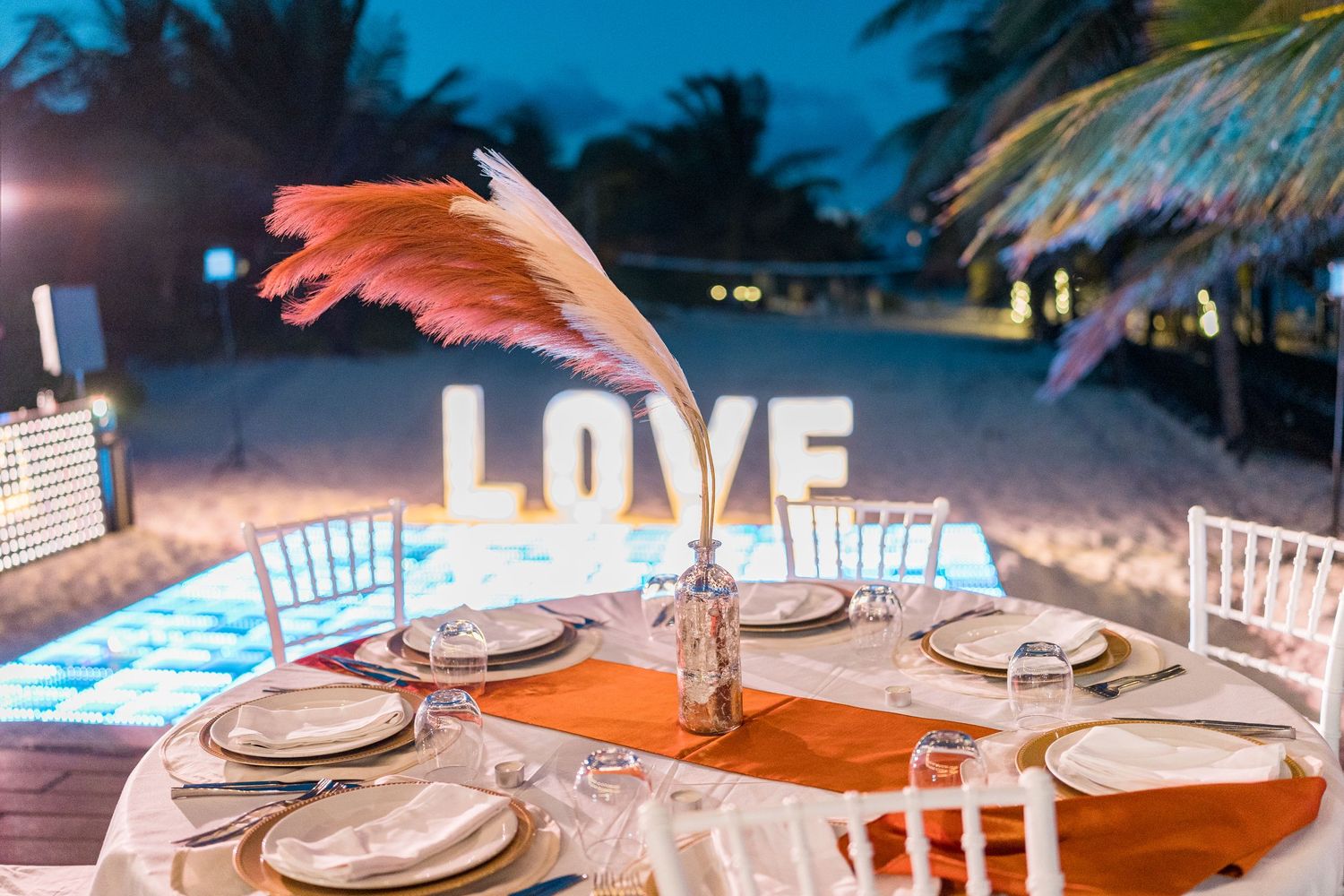 Beach wedding reception table with orange feather centerpiece and illuminated LOVE sign in background.