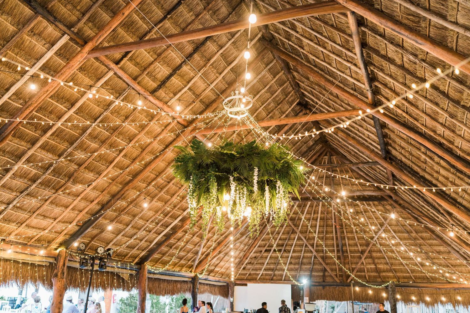 Rustic palapa wedding venue with string lights and lush green floral chandelier hanging from thatched roof.
