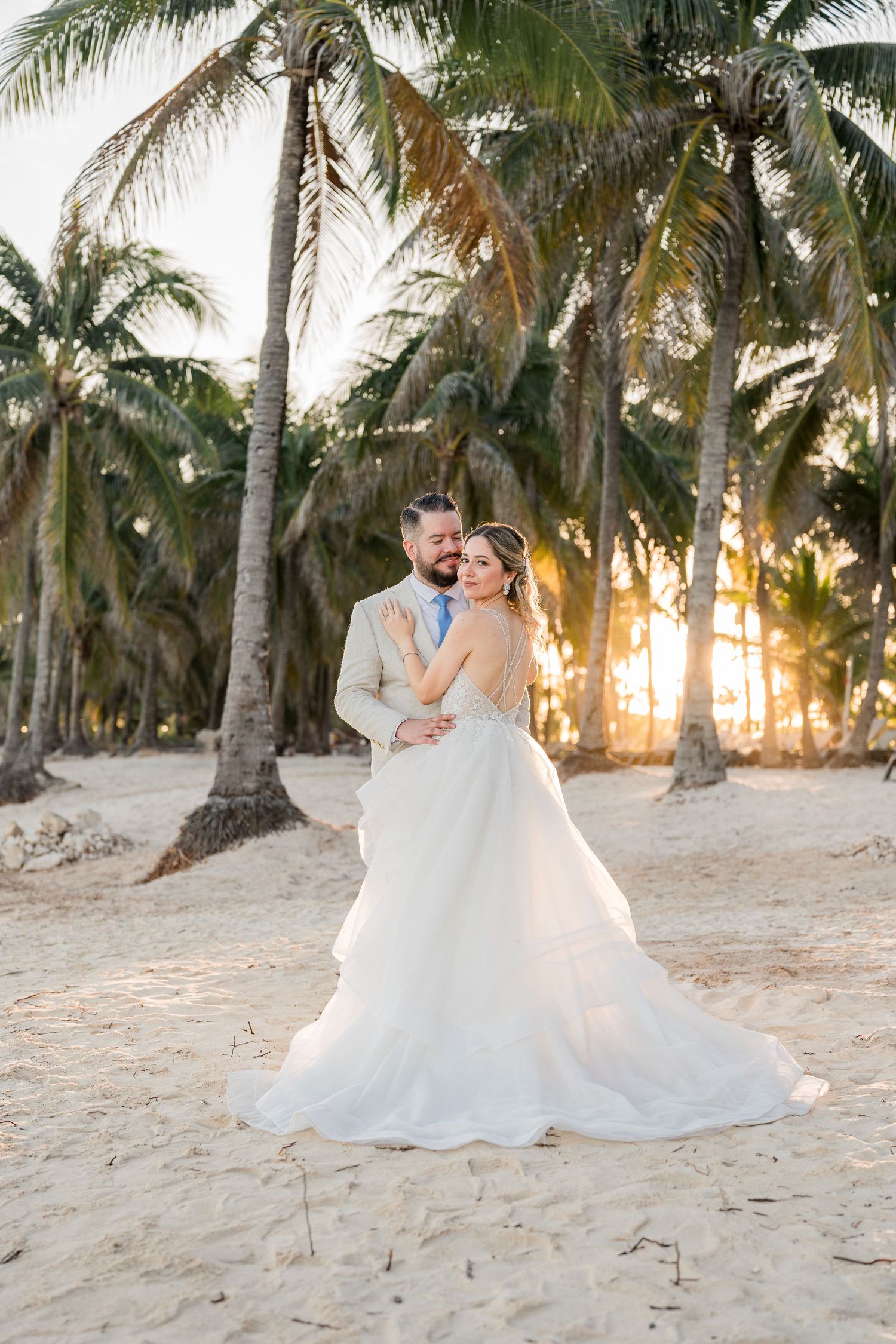 Bride and groom embrace on a sandy beach at sunset surrounded by tall tropical palm trees.