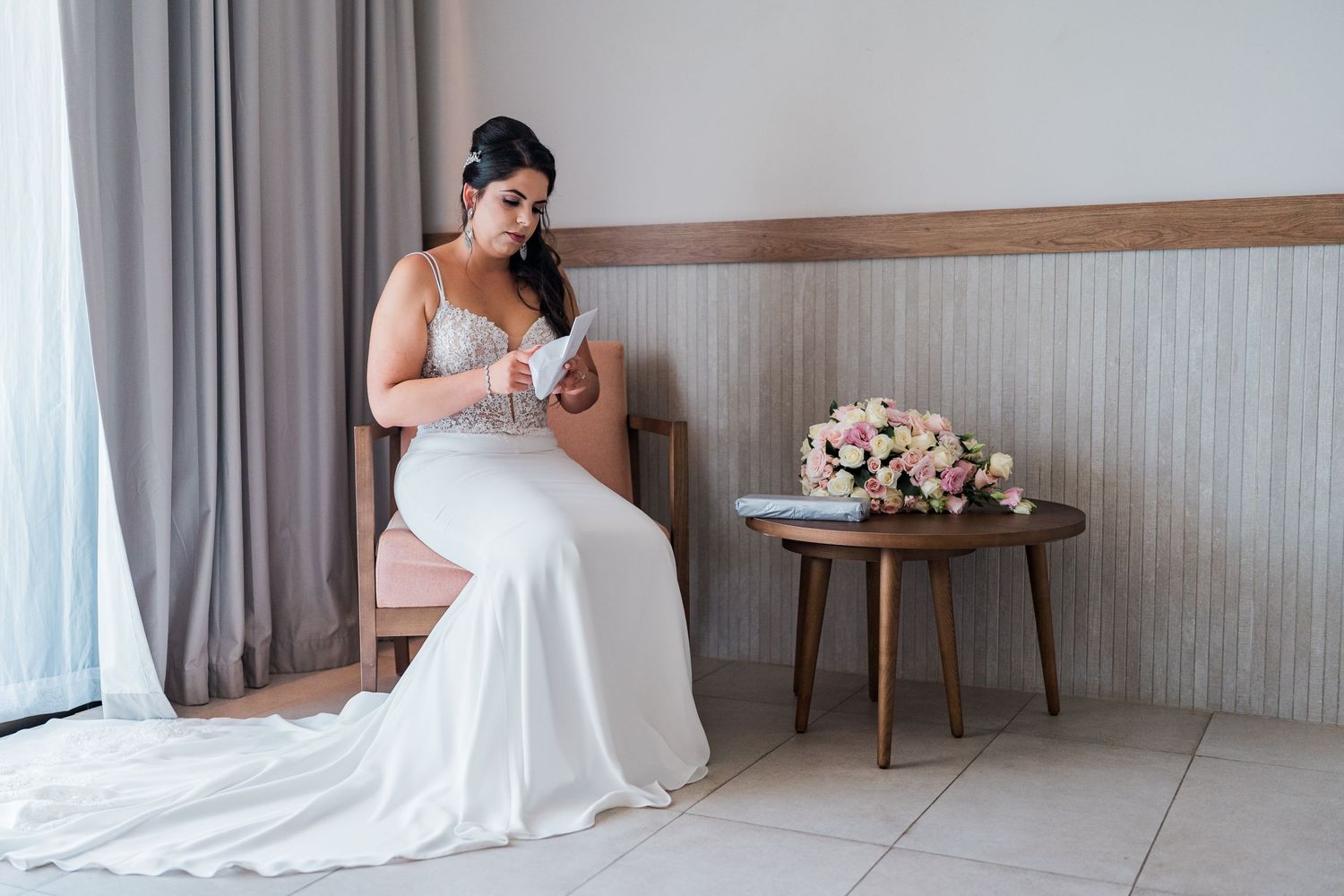 Bride in white gown sitting in chair reading note beside pink floral bouquet.