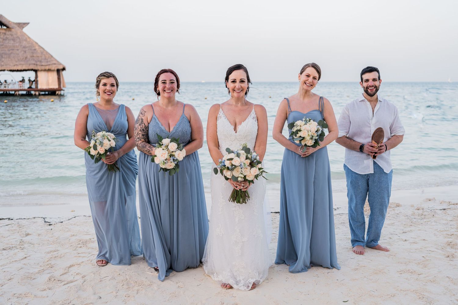 Bride in white gown with bridesmaids in blue dresses and groomsman on beach.