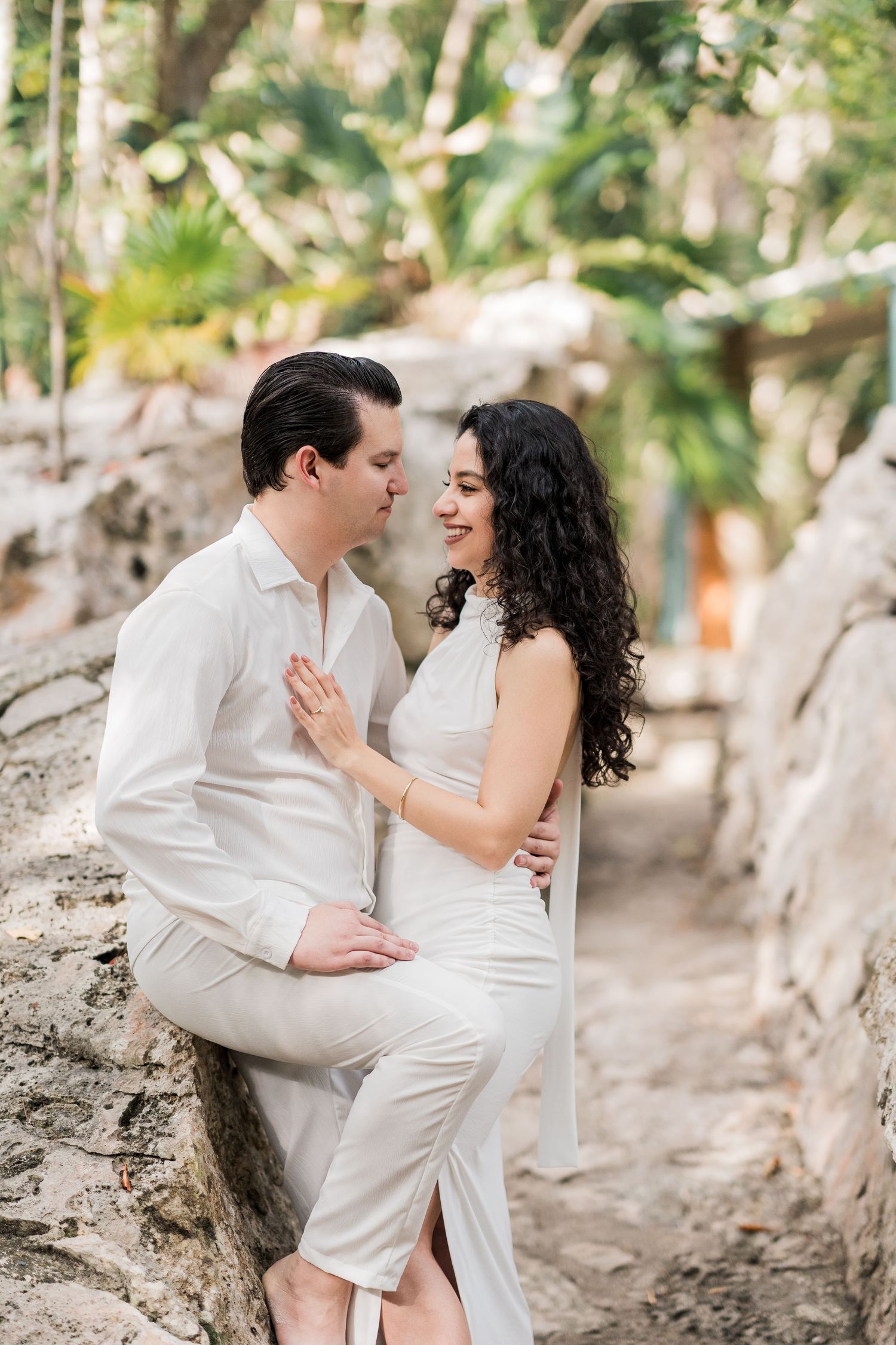 A couple in white outfits share a romantic moment on stone ruins surrounded by tropical greenery.