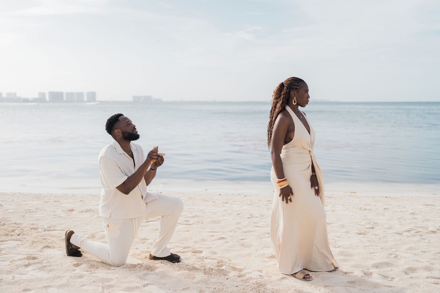 Man proposing on one knee to woman in white dress on a sunny beach with ocean in background.