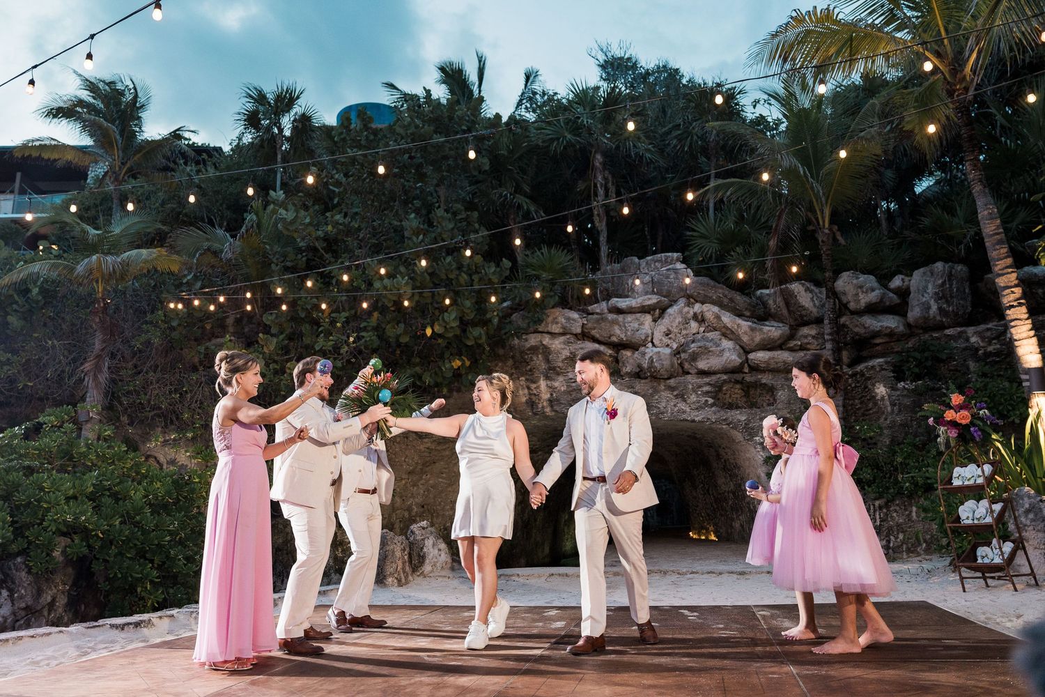 Wedding party dancing outdoors at tropical venue with string lights, rocky backdrop, and palm trees at dusk.
