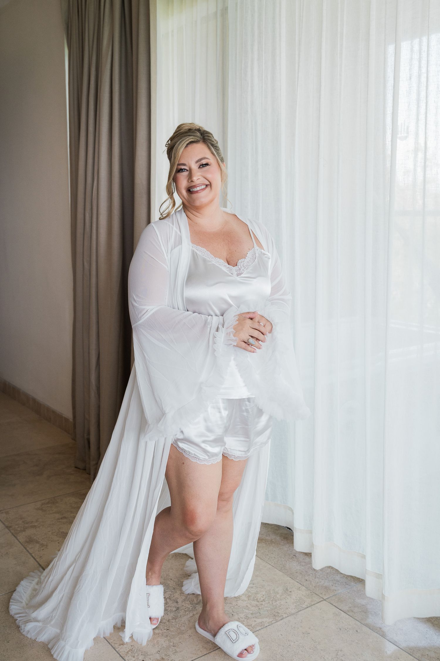 Smiling bride in white satin shorts set and sheer robe with bridal slippers posing by sheer curtains.