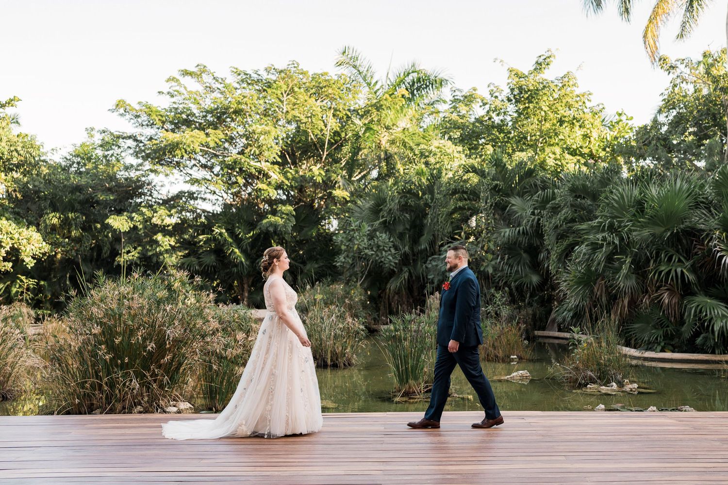 Secrets Akumal Wedding - Bride and groom walking on wooden deck surrounded by tropical greenery and pond at outdoor wedding venue.