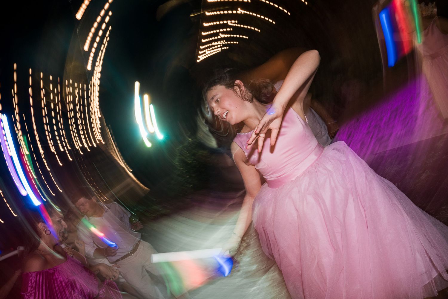 A young woman in a pink tulle dress dances joyfully at a party, surrounded by swirling colorful lights.