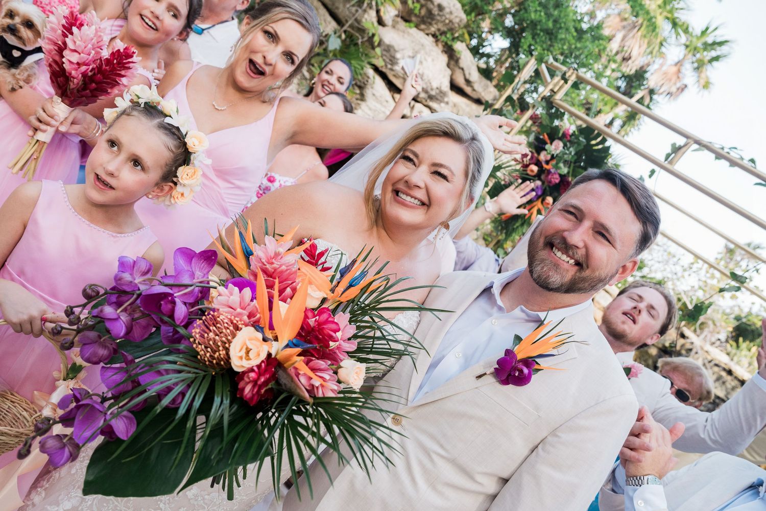 Bride and groom smile with bridesmaids in pink holding tropical bouquet with birds of paradise at outdoor wedding.