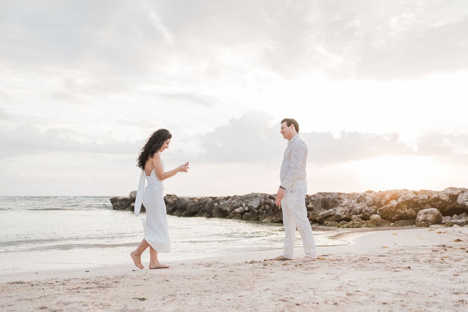 Couple in white outfits sharing a romantic moment on a sandy beach at sunset with rocky shoreline.