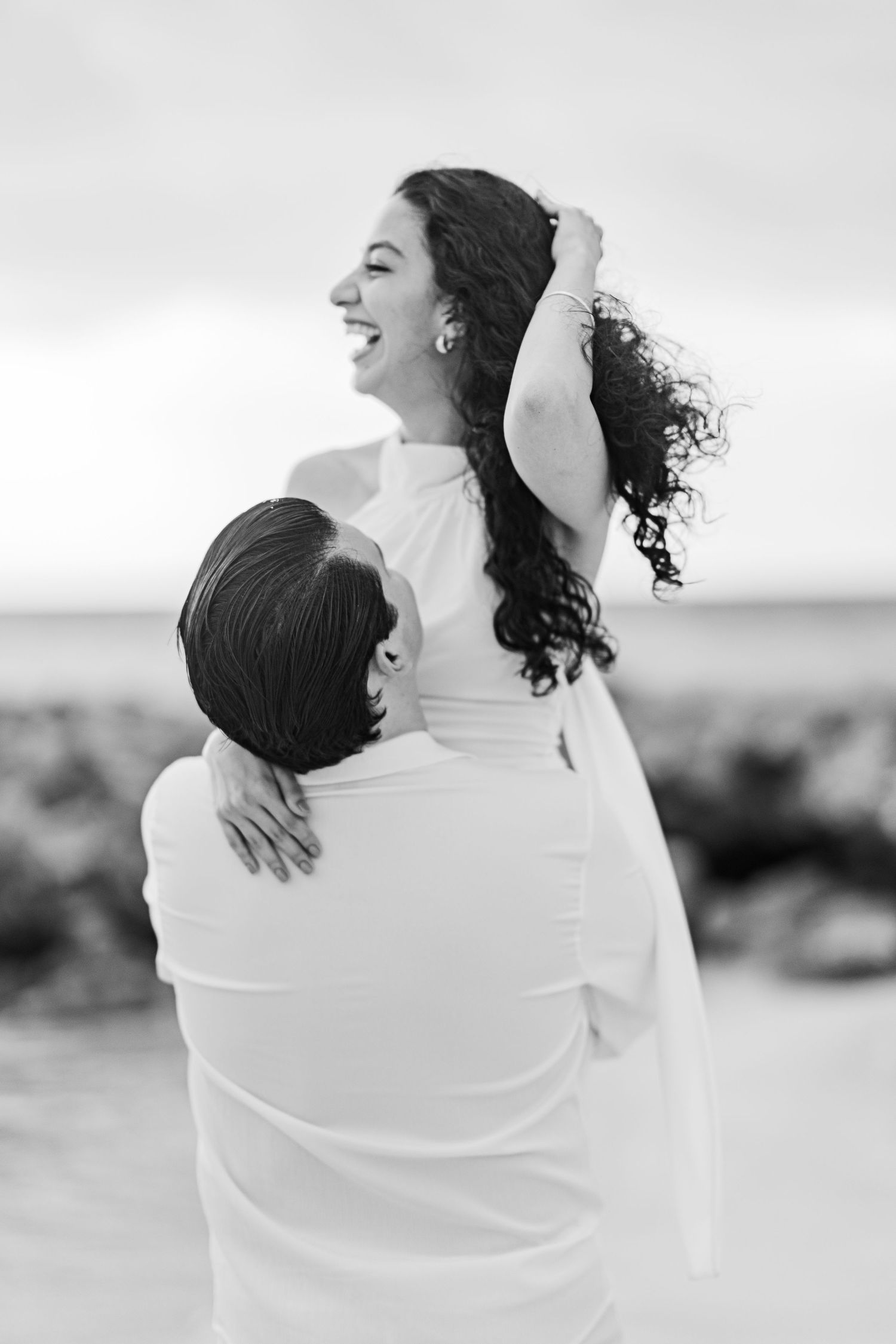 Black and white beach wedding photo of groom lifting smiling bride in white dress, her curly hair blowing in the wind.