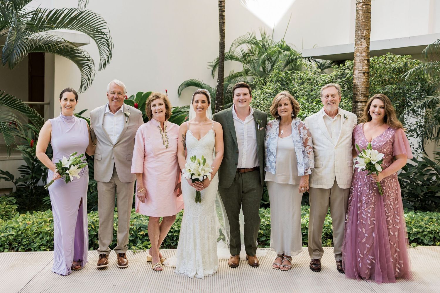 Wedding party of eight posing outdoors, featuring bride in white gown, groom in olive suit, and guests in pastel formal attire.