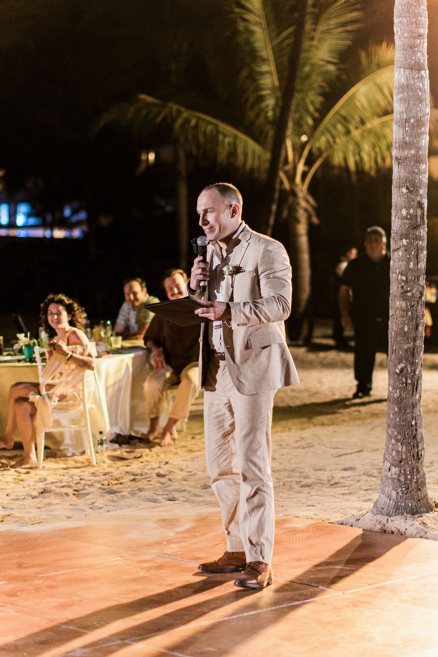 Man in tan suit giving speech at outdoor beach wedding reception at night with palm trees.