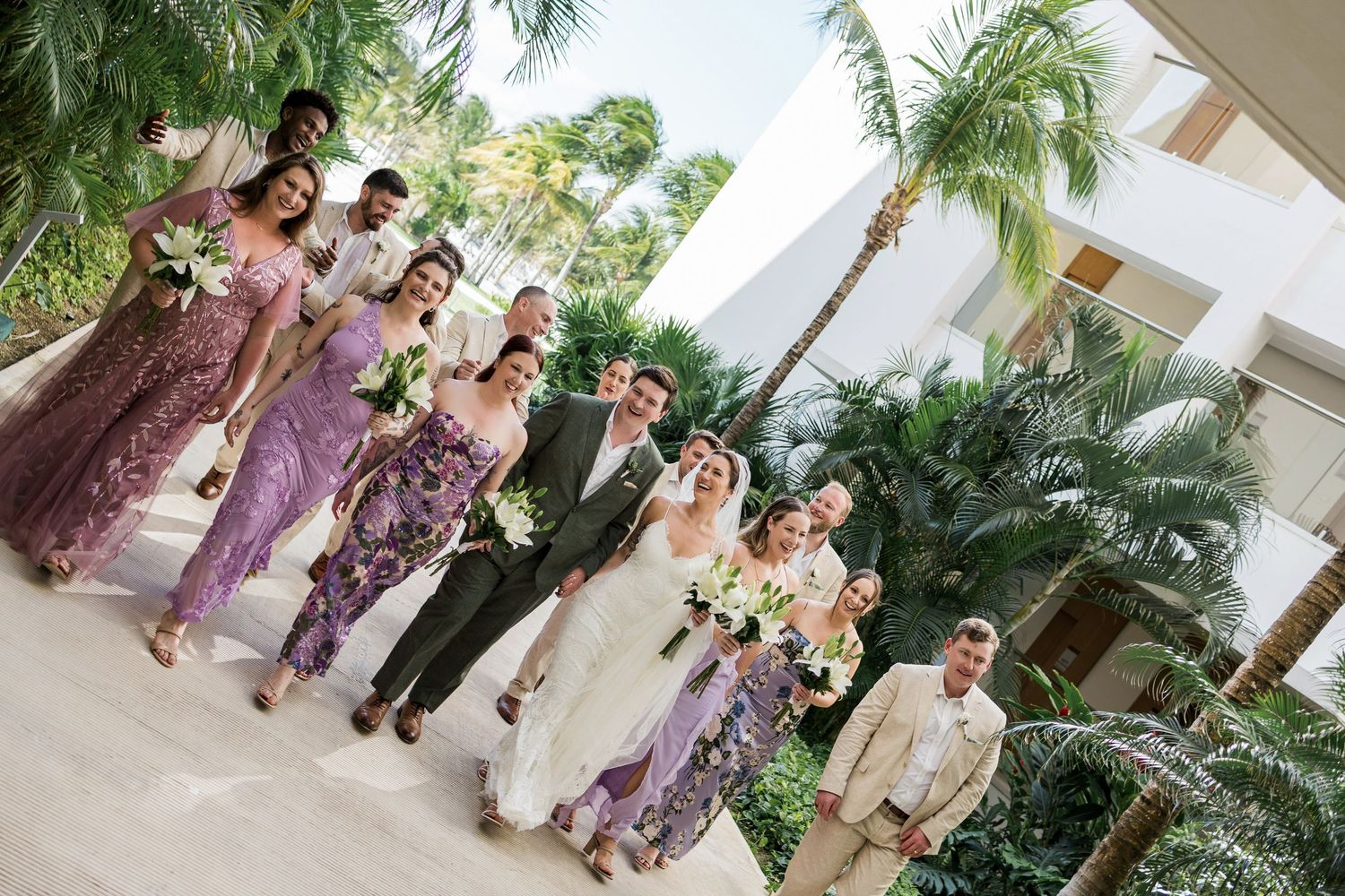 Wedding party walks together outdoors, bridesmaids in purple lace dresses, groomsmen in tan suits, bride in white gown, tropical palm trees.
