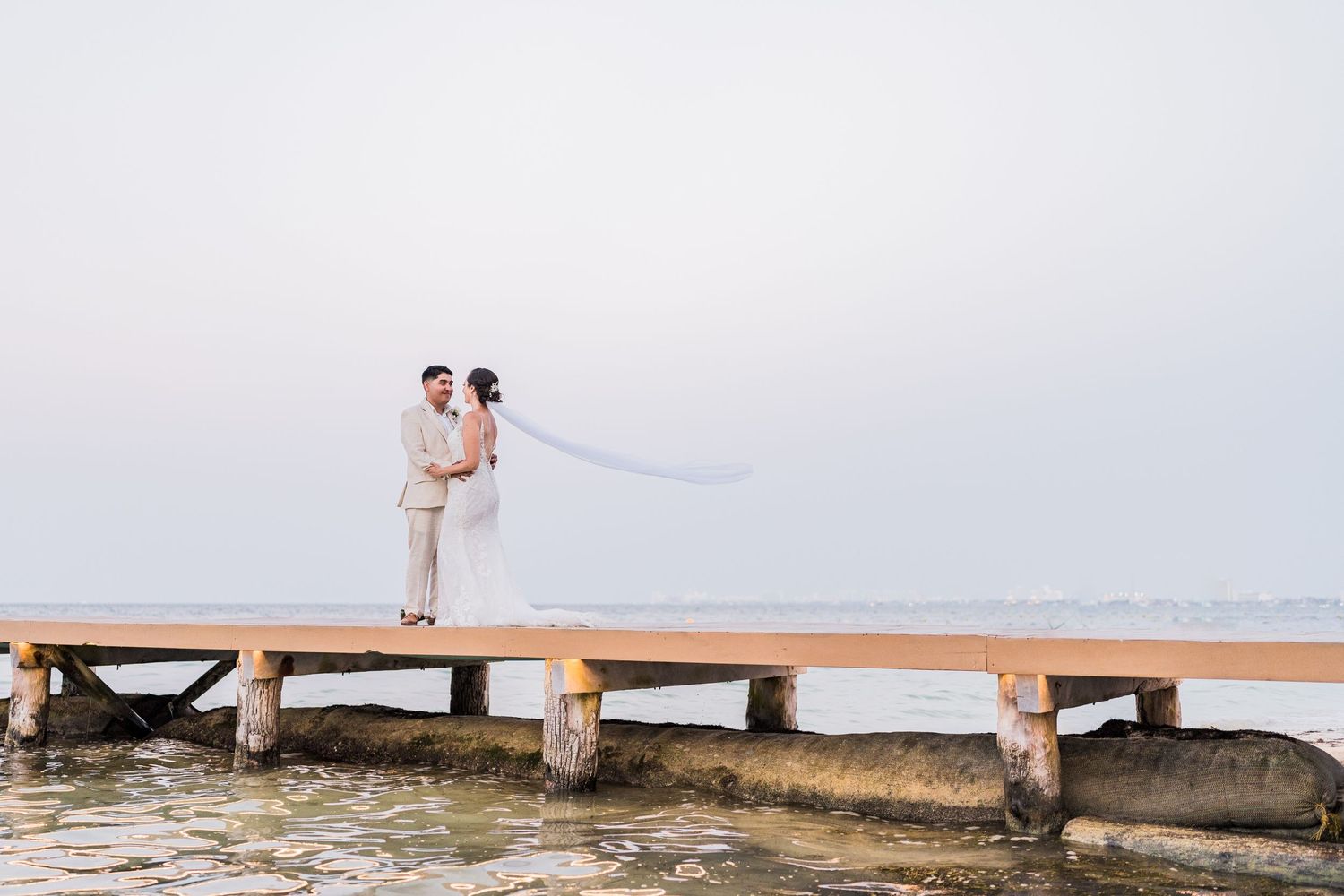 Bride and groom share a romantic moment on a wooden pier over the ocean at sunset.