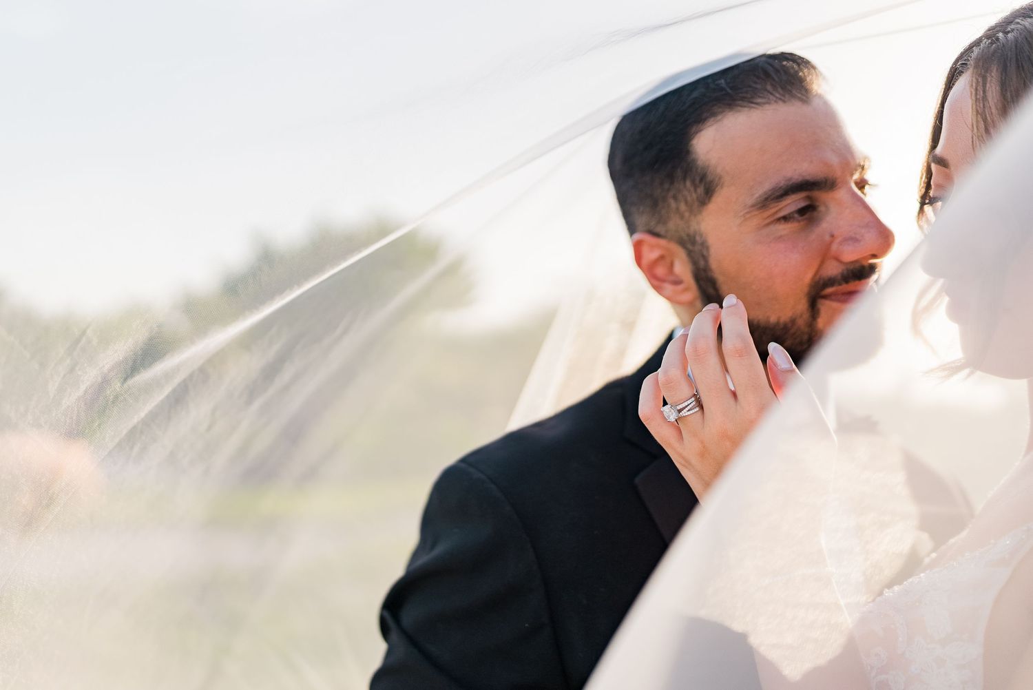 Groom tenderly holds bride's face under her flowing wedding veil in golden light.