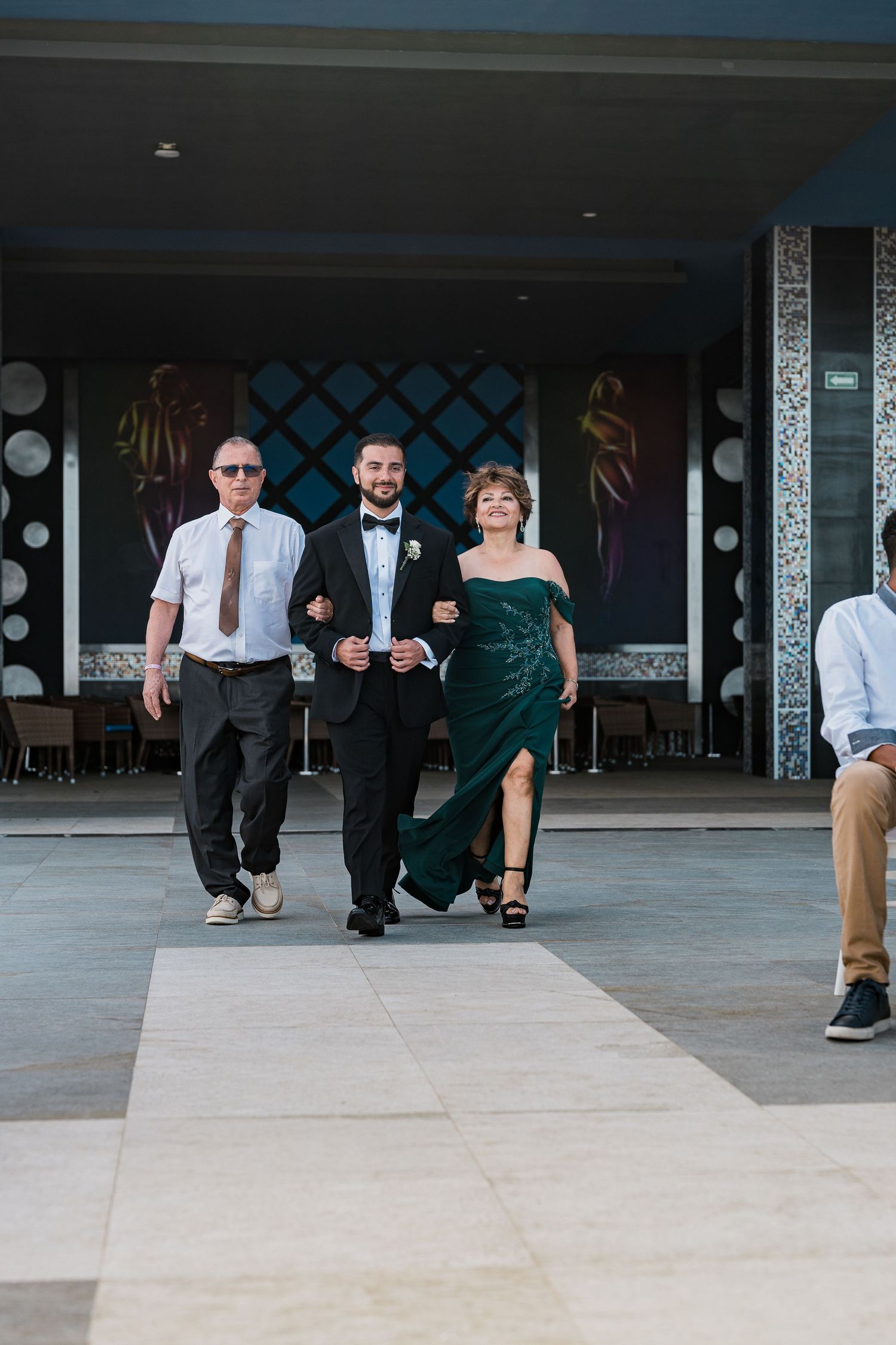 Groom walks down aisle escorted by parents at Mexico beach wedding ceremony.