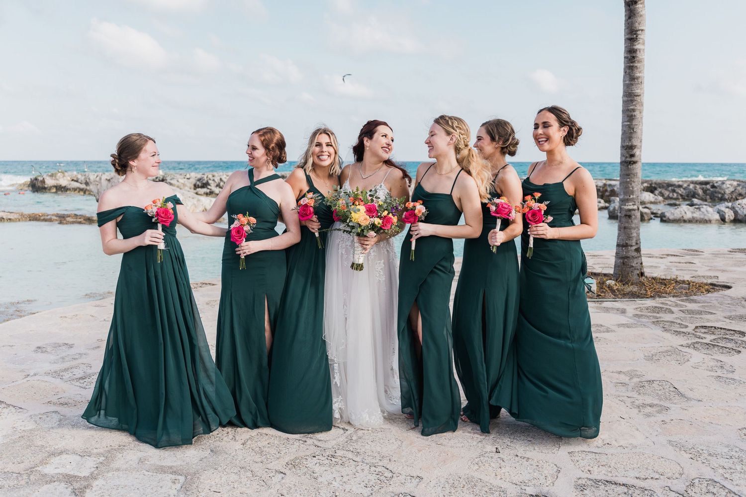 Bride poses with six bridesmaids in emerald green gowns holding pink bouquets by the ocean.