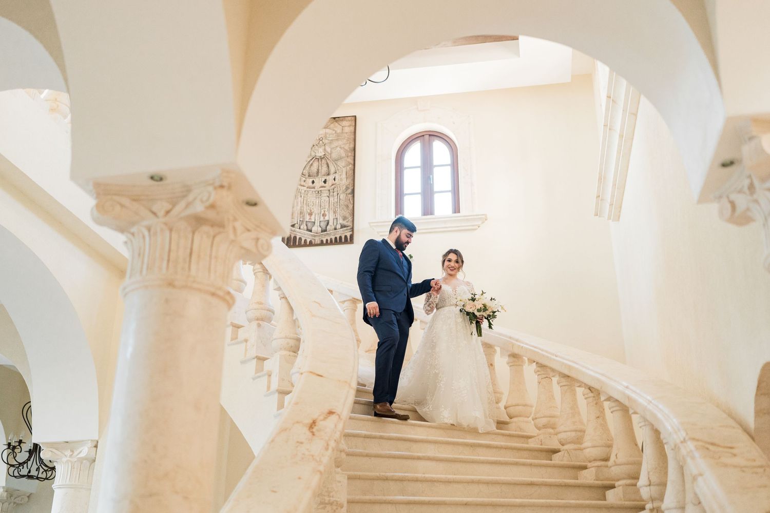 Bride and groom descend ornate marble staircase as bride holds white floral bouquet.