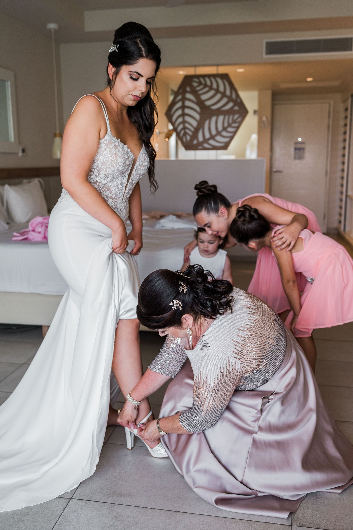 Bride getting shoes fastened by bridesmaid while flower girls watch in elegant hotel room.