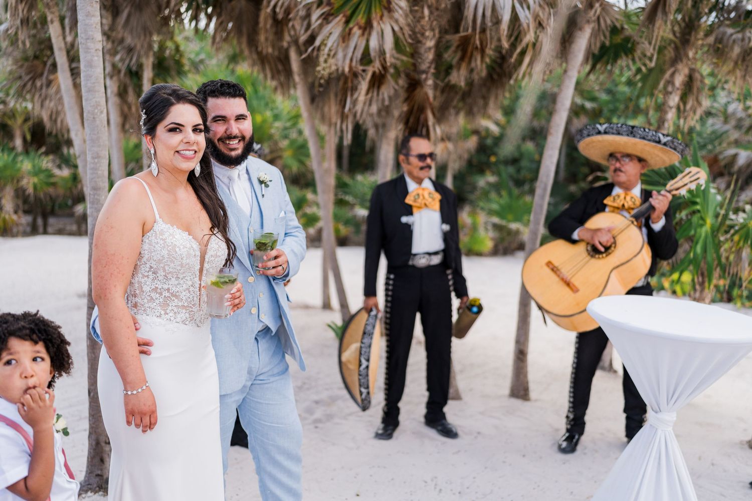 Bride and groom smiling together on a beach while a mariachi band performs nearby.