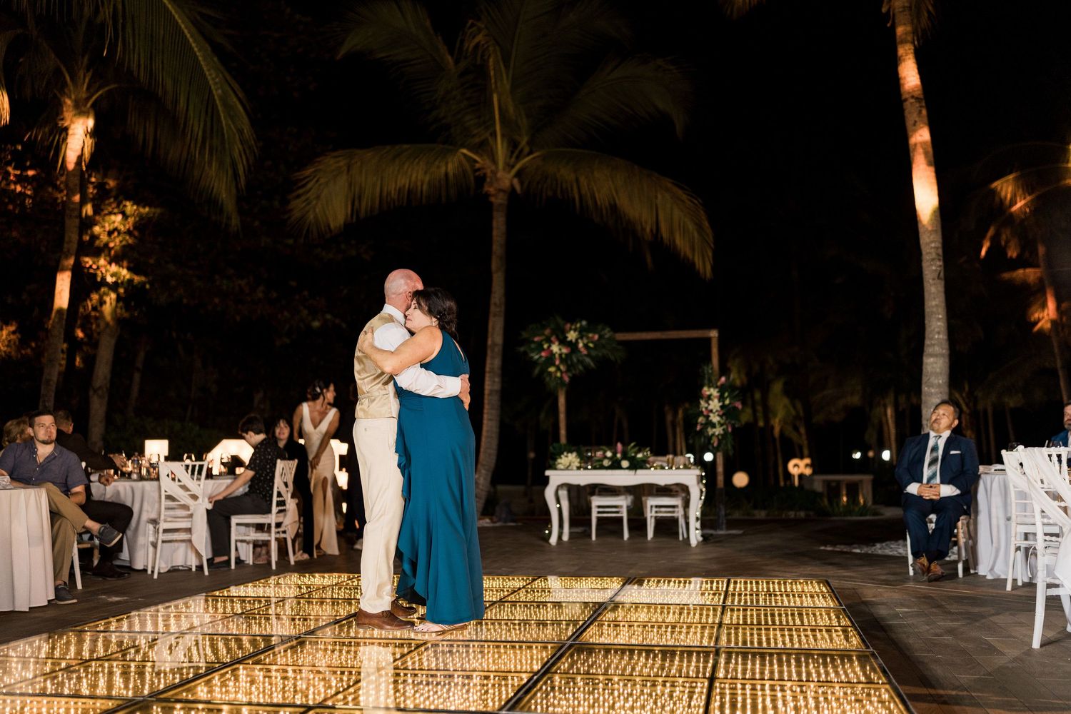 Couple shares first dance on illuminated floor at outdoor tropical wedding reception at night with palm trees.