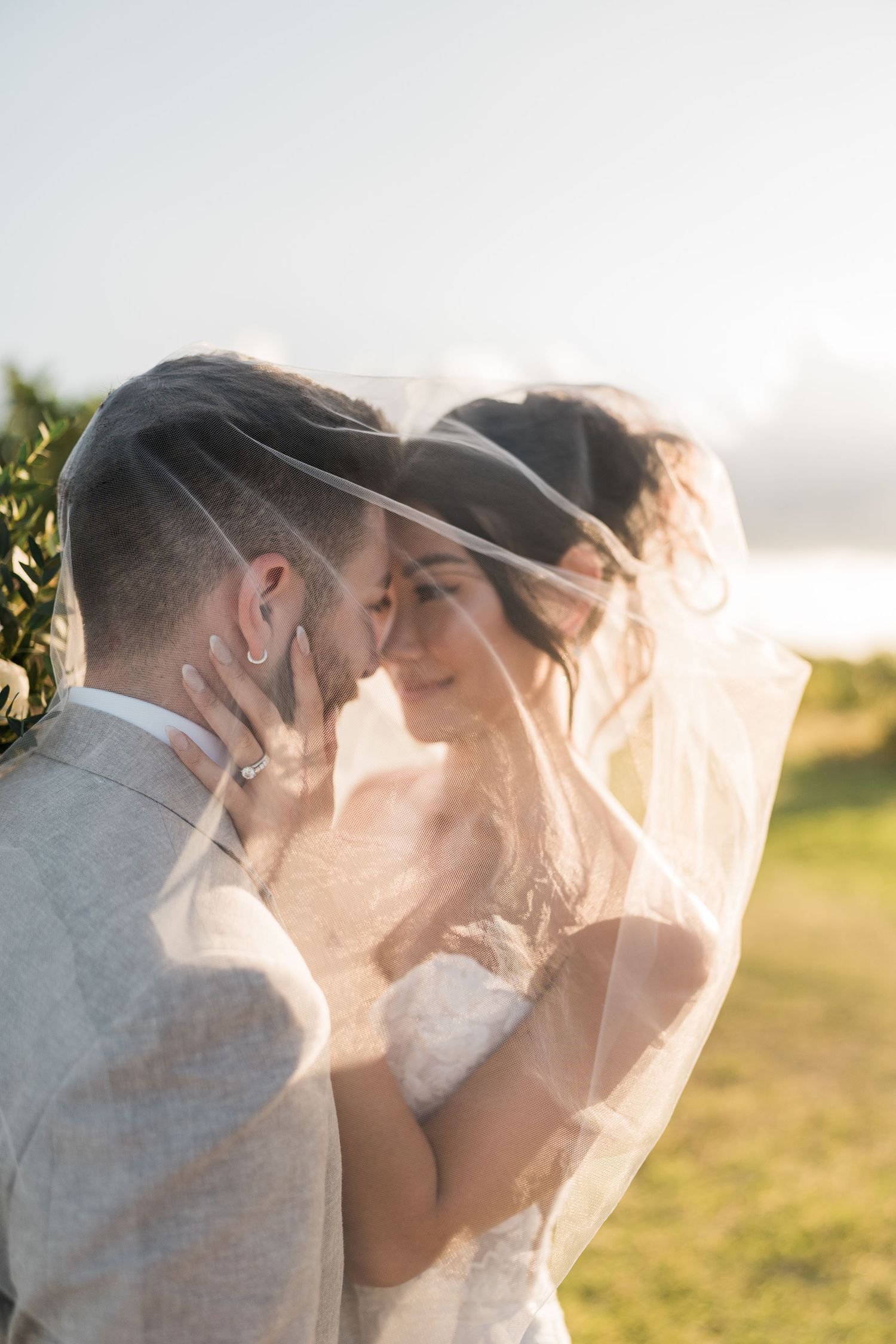 Bride and groom share intimate moment under wedding veil at golden hour outdoors.