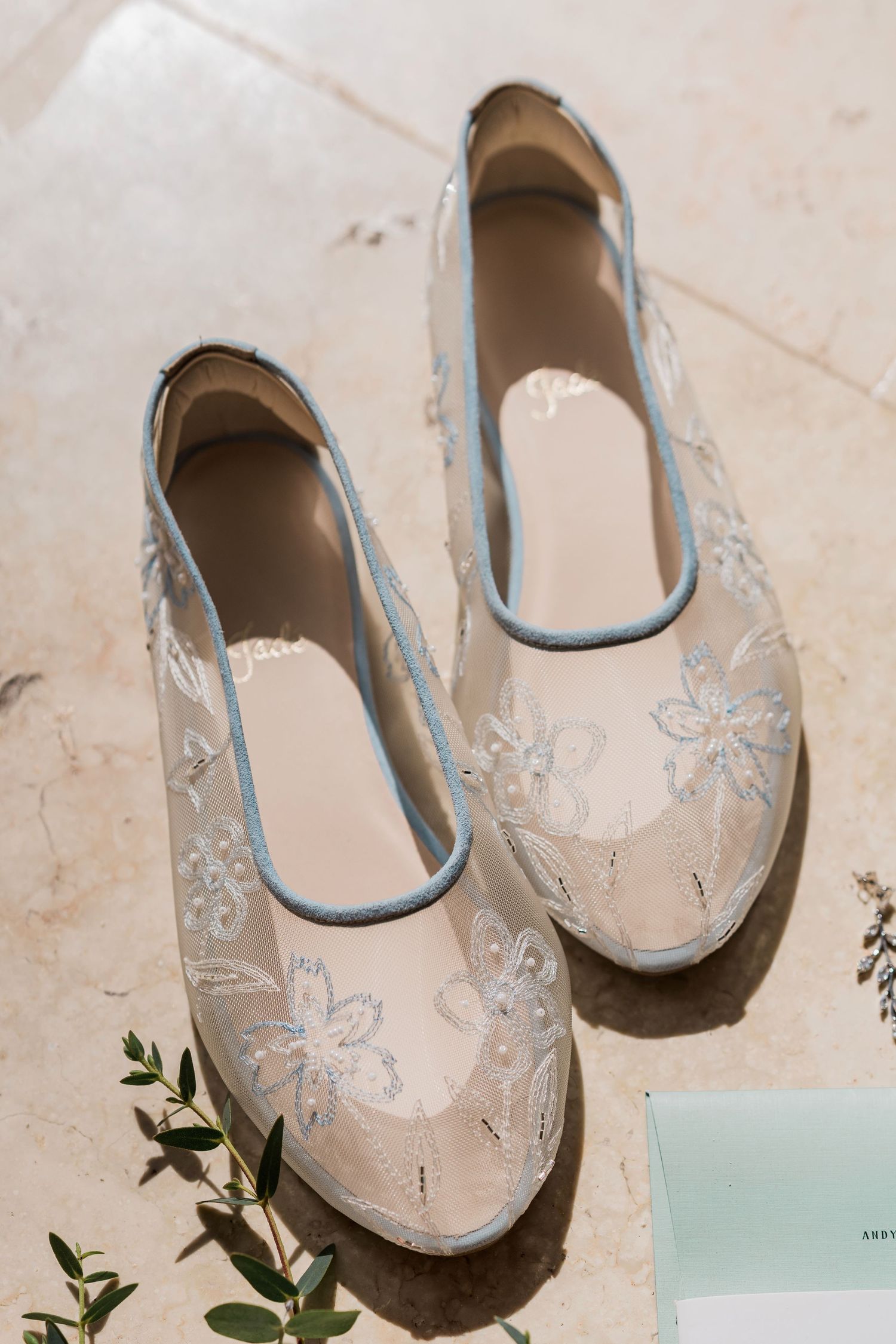 Elegant sheer lace bridal flats with blue trim and floral embroidery displayed on marble surface.