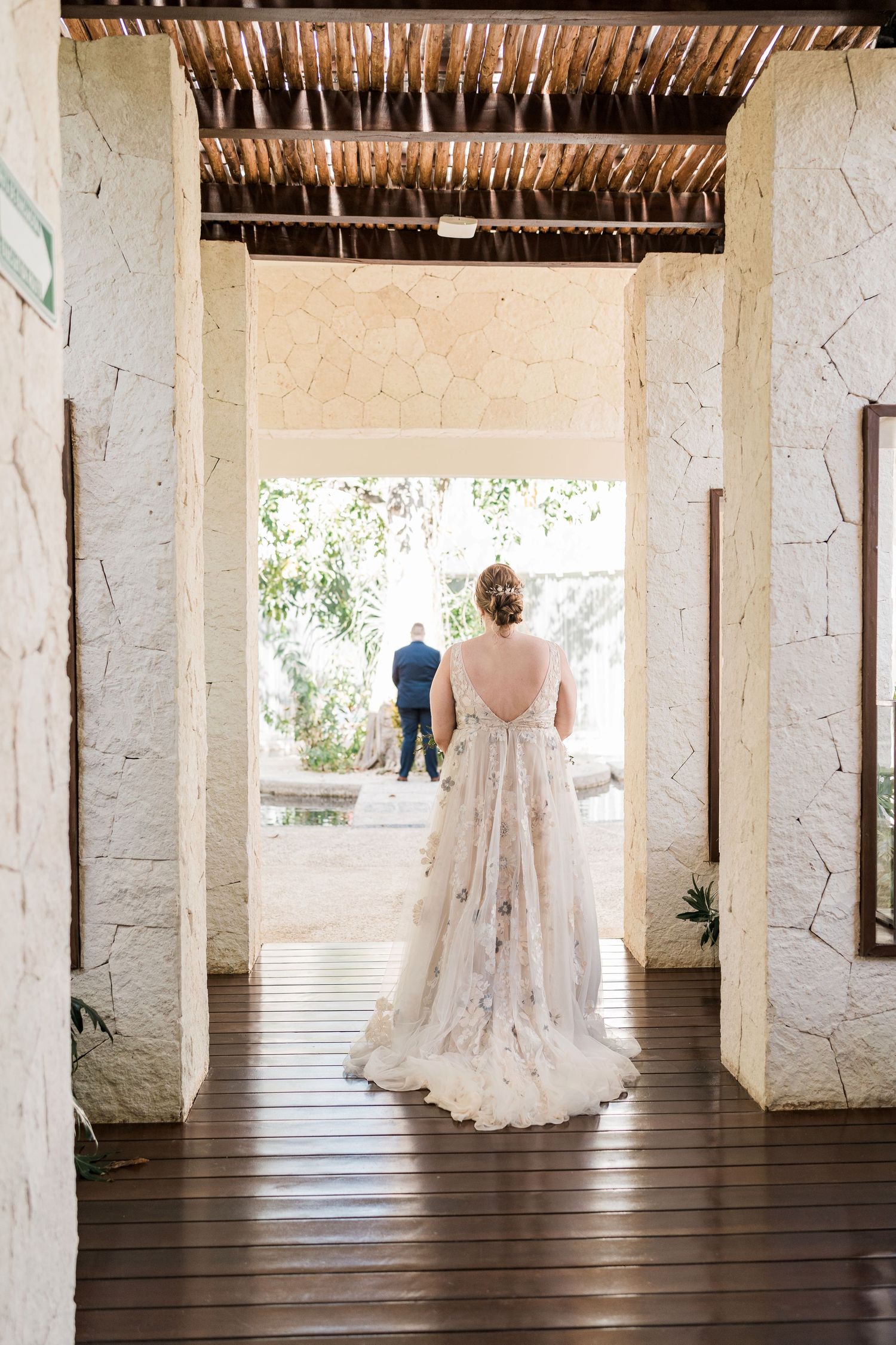 Secrets Akumal Wedding - Bride in a floral gown walks toward groom in a tropical venue with stone walls and wooden ceiling.