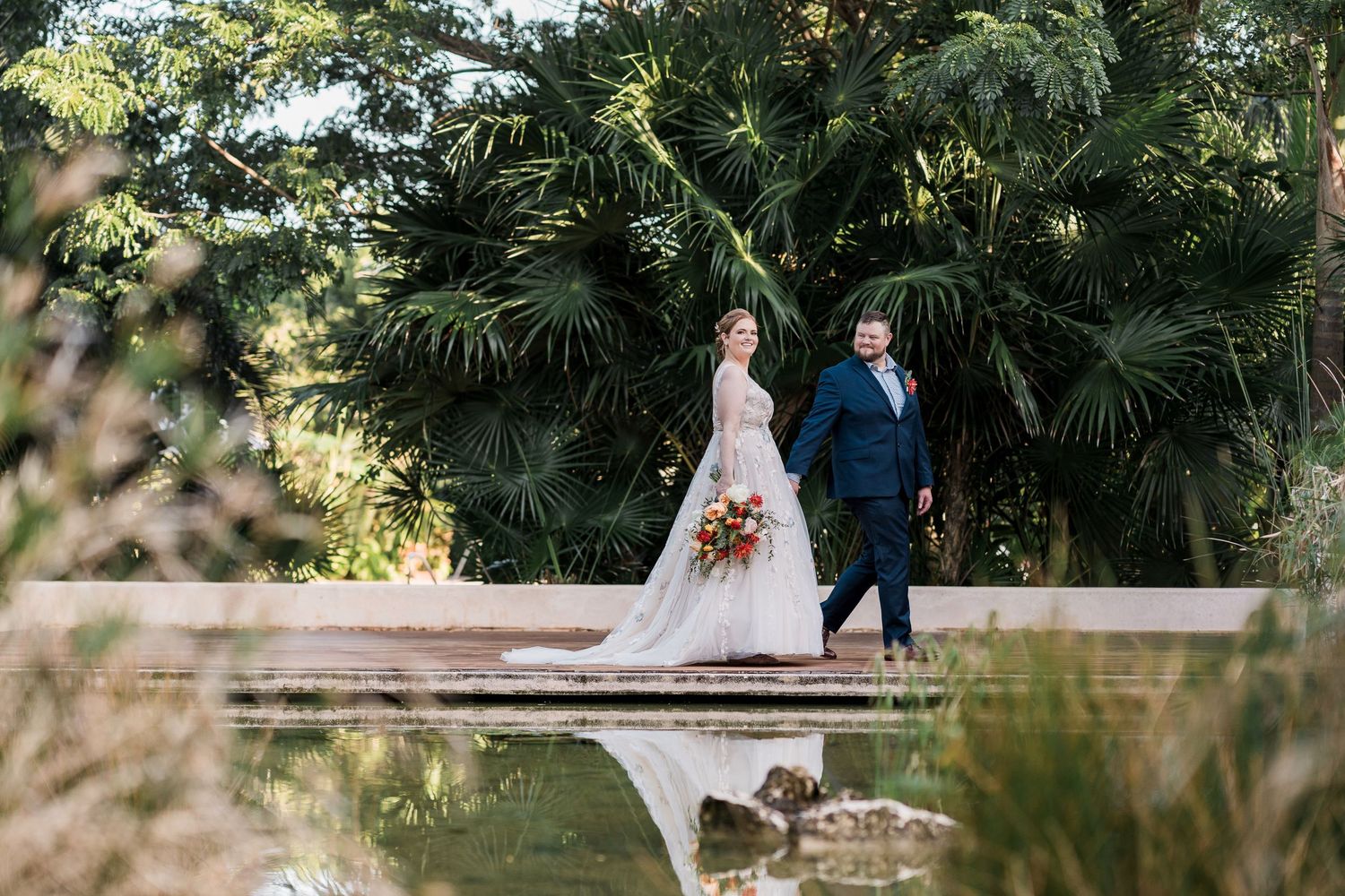 Secrets Akumal Wedding - Bride and groom walking hand in hand beside a reflective pond surrounded by lush tropical greenery.