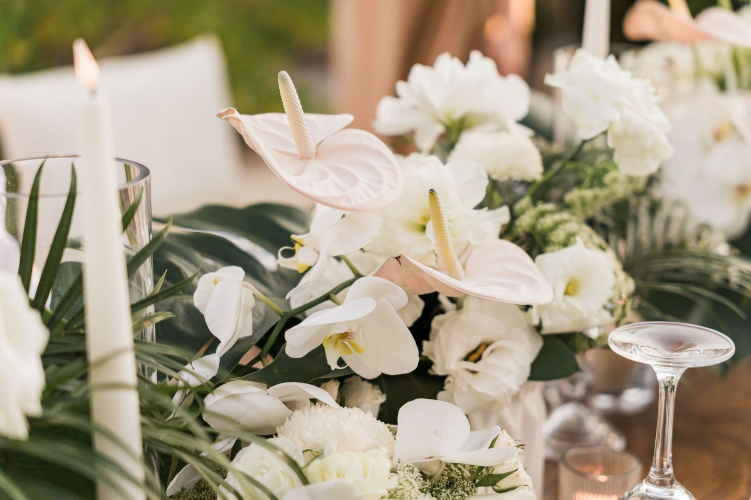 Xcaret Arte Wedding - Elegant white floral centerpiece with anthuriums, lilies, and tropical leaves beside candles and glassware.