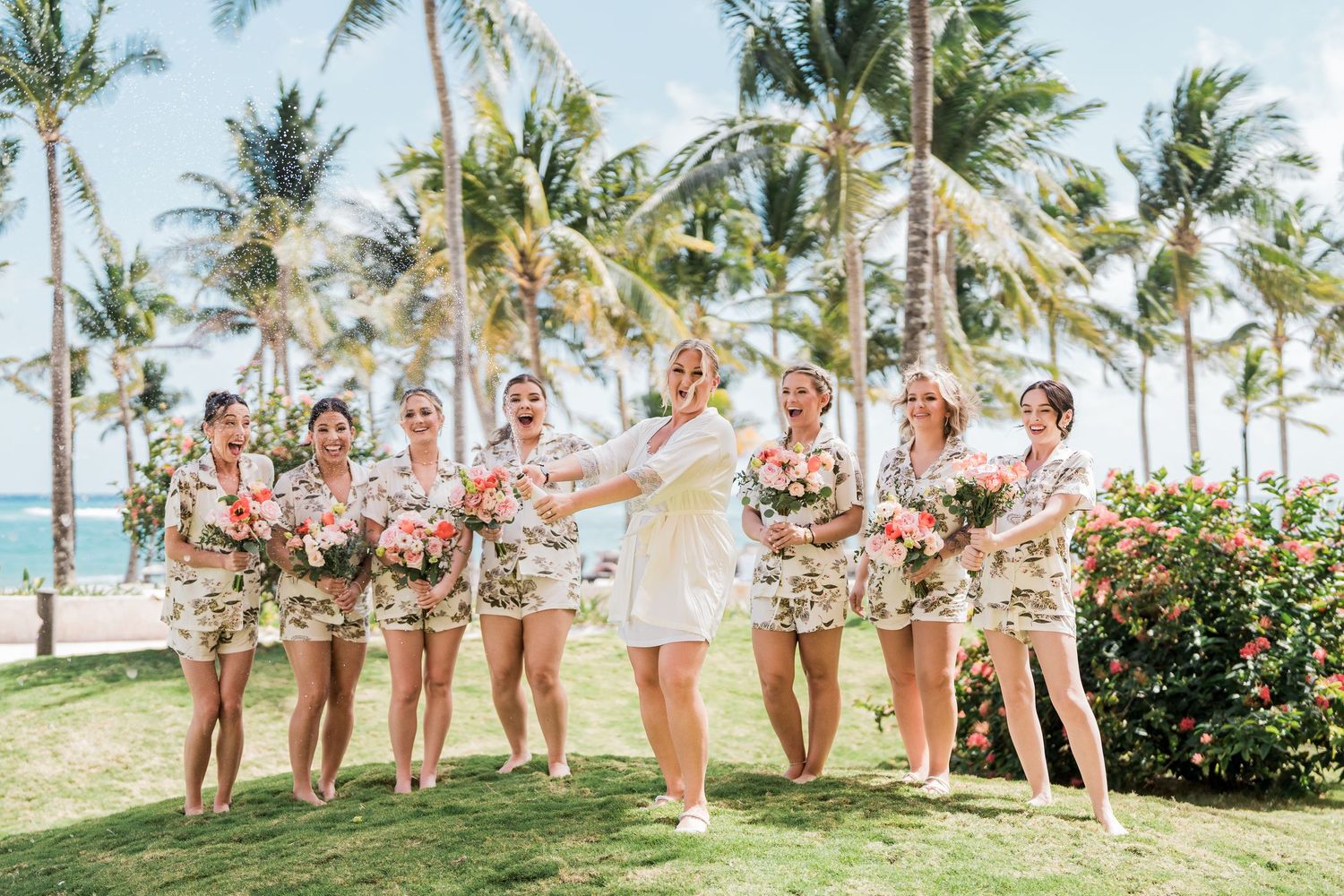 Bride in white robe celebrates with bridesmaids in floral matching sets on tropical beach lawn.