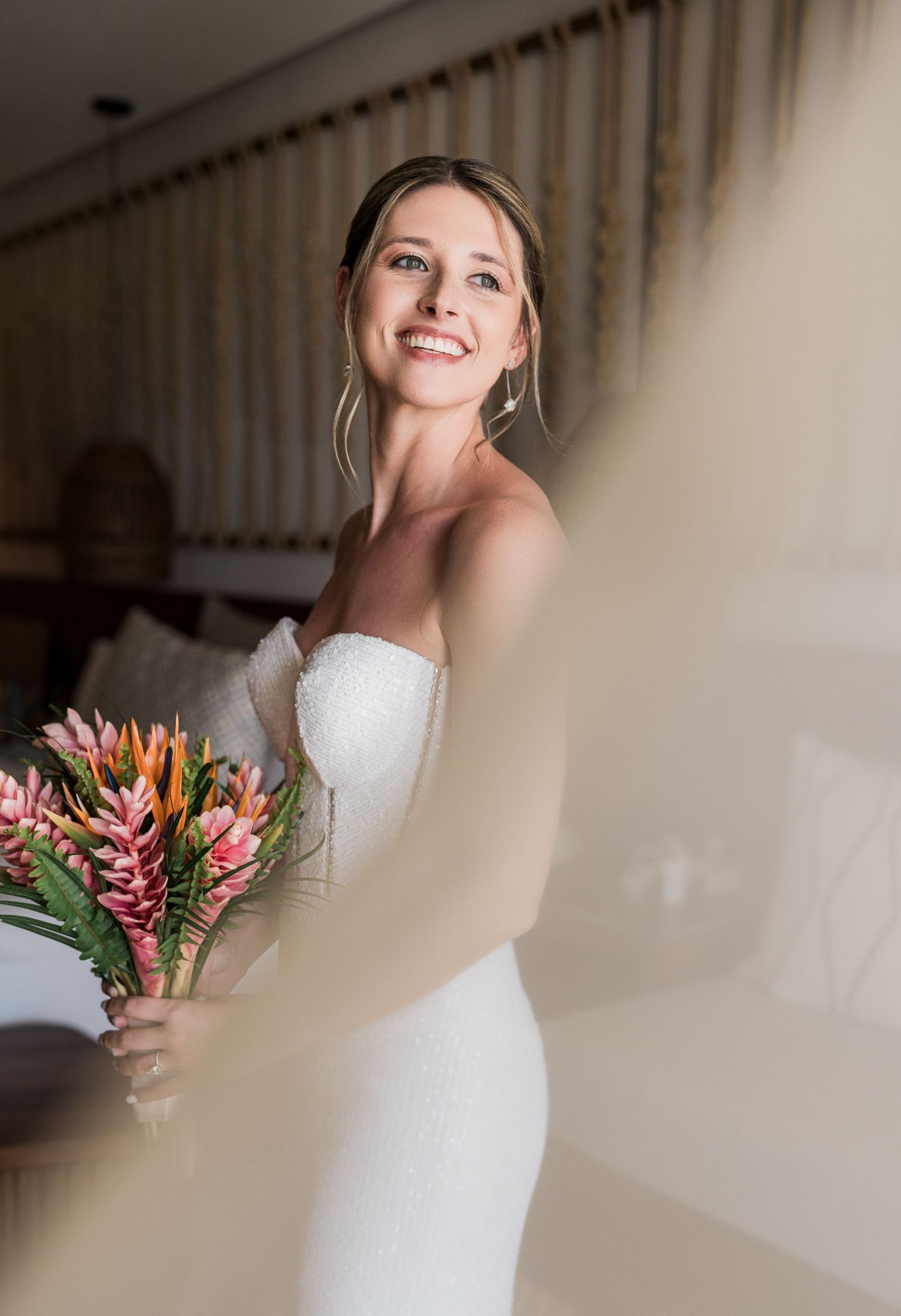 Smiling bride in strapless white gown holds tropical pink bouquet, standing in soft natural light.