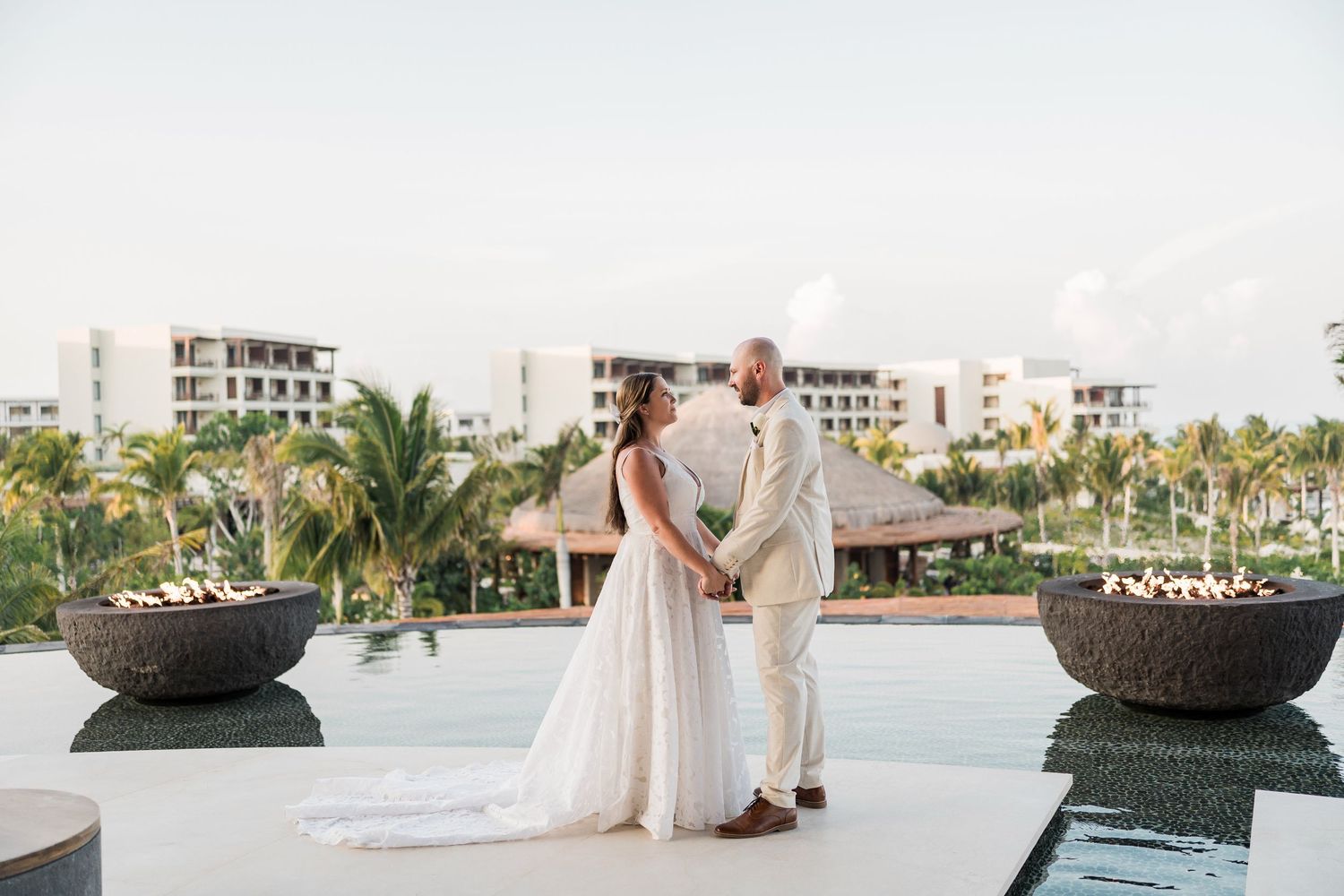 Bride and groom hold hands on a rooftop terrace with fire bowls, pool, and tropical resort backdrop at sunset.