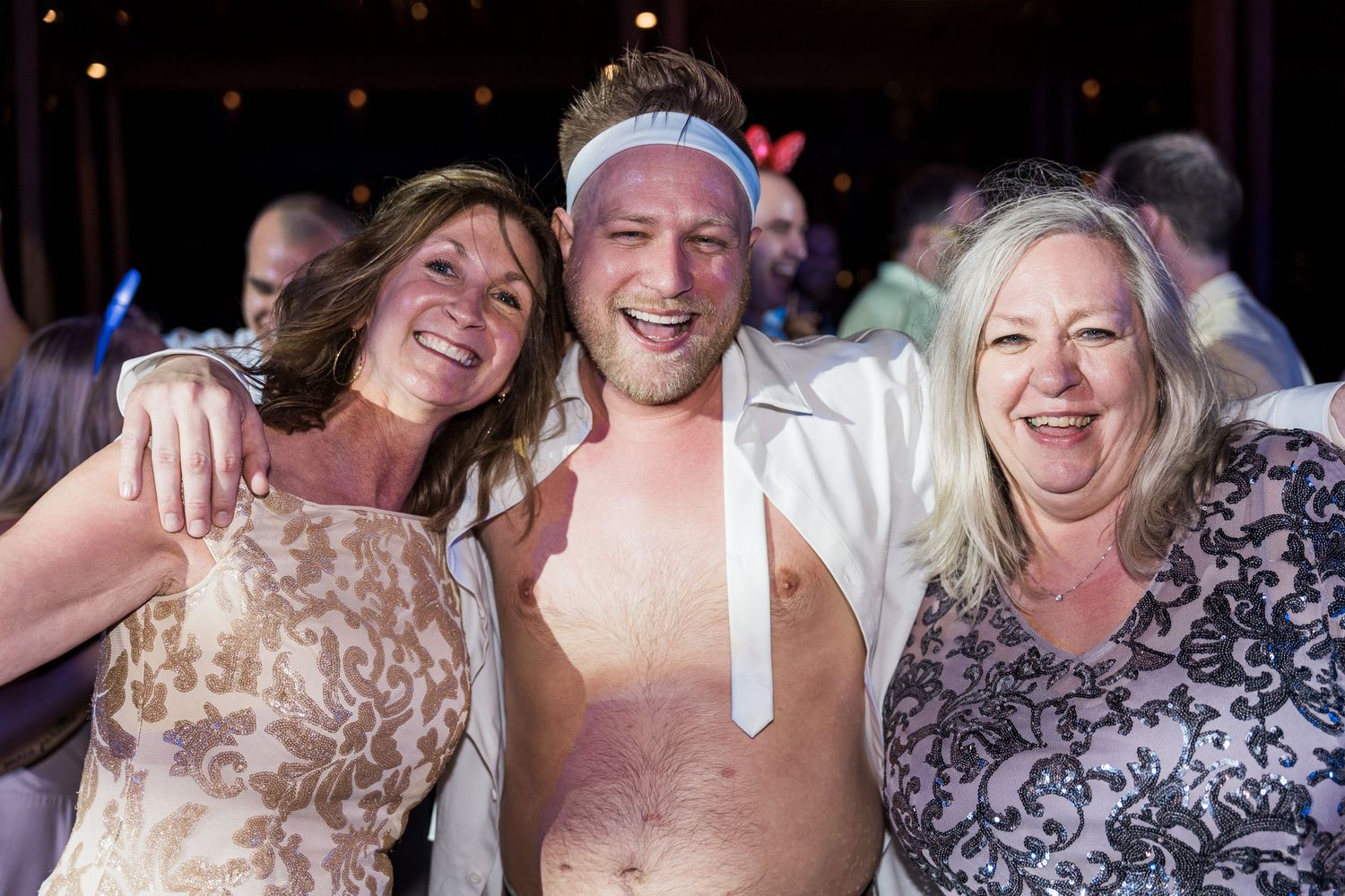 Three adults smiling and posing together at a lively party event, with the man in the center wearing a headband.