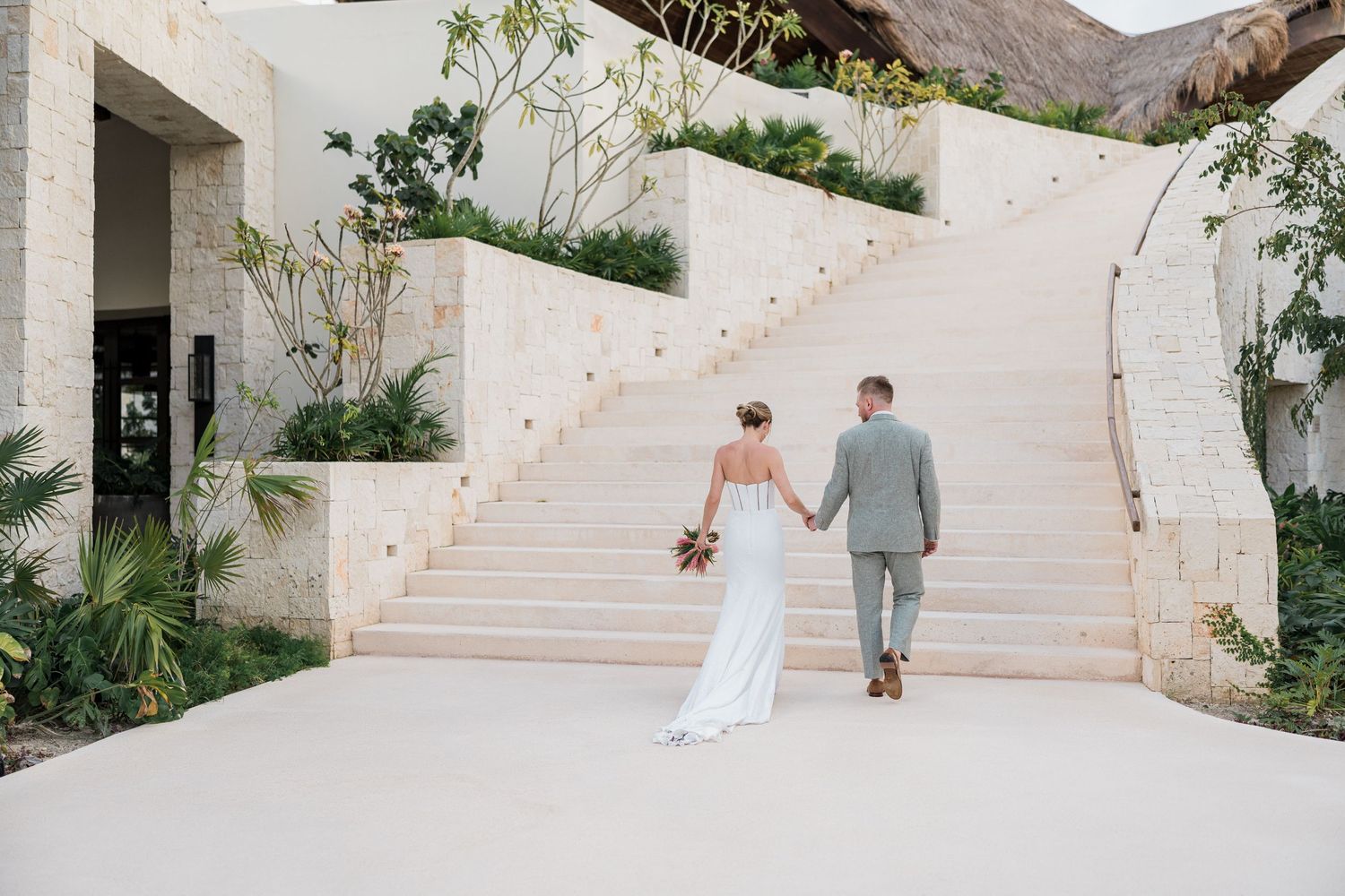 Bride and groom holding hands walking toward grand white staircase at luxury outdoor wedding venue.