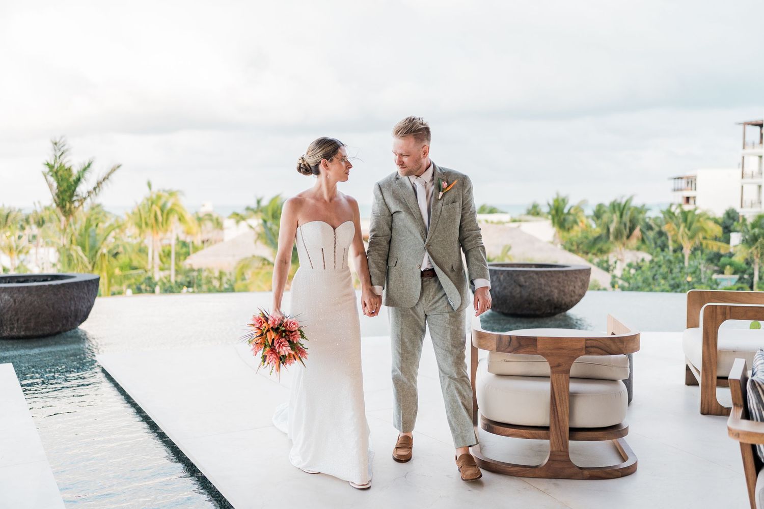 Bride and groom walking hand in hand on a luxury resort terrace with tropical views and a pool.
