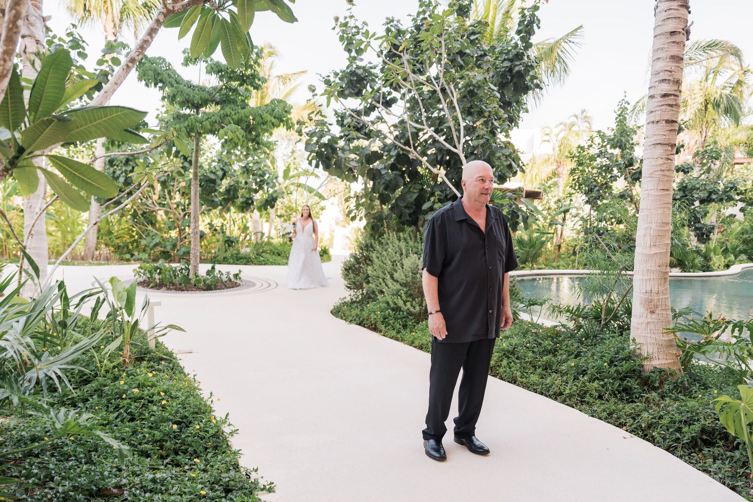 Man in black attire walks a tropical garden path as a bride in white dress stands in the background.