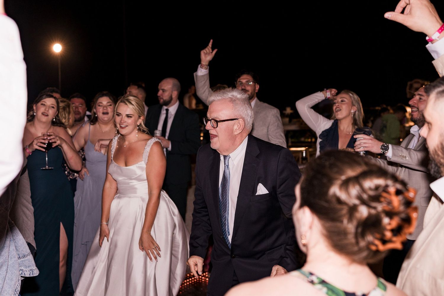 Bride in white gown dances joyfully with older gentleman in suit at elegant outdoor wedding reception at night.