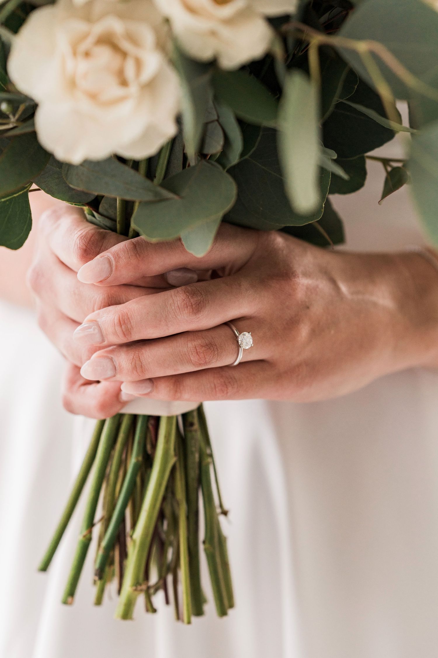 Bride holding a white rose and eucalyptus wedding bouquet, wearing a diamond engagement ring on her finger.