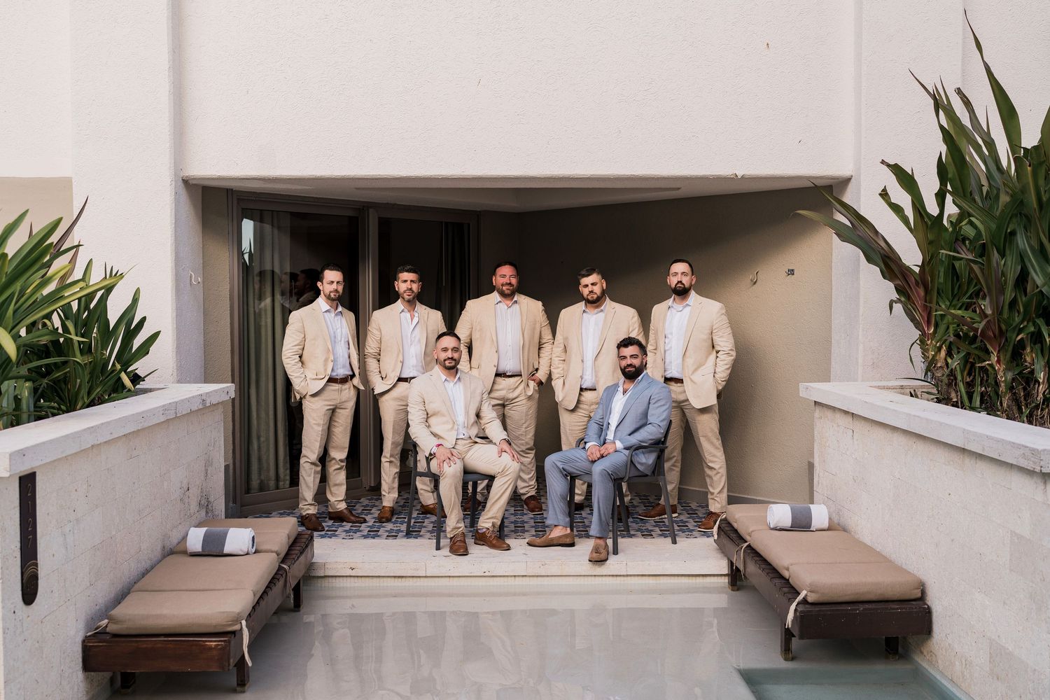 Groom and groomsmen in tan suits posing on a modern outdoor terrace with lounge chairs at a wedding venue.
