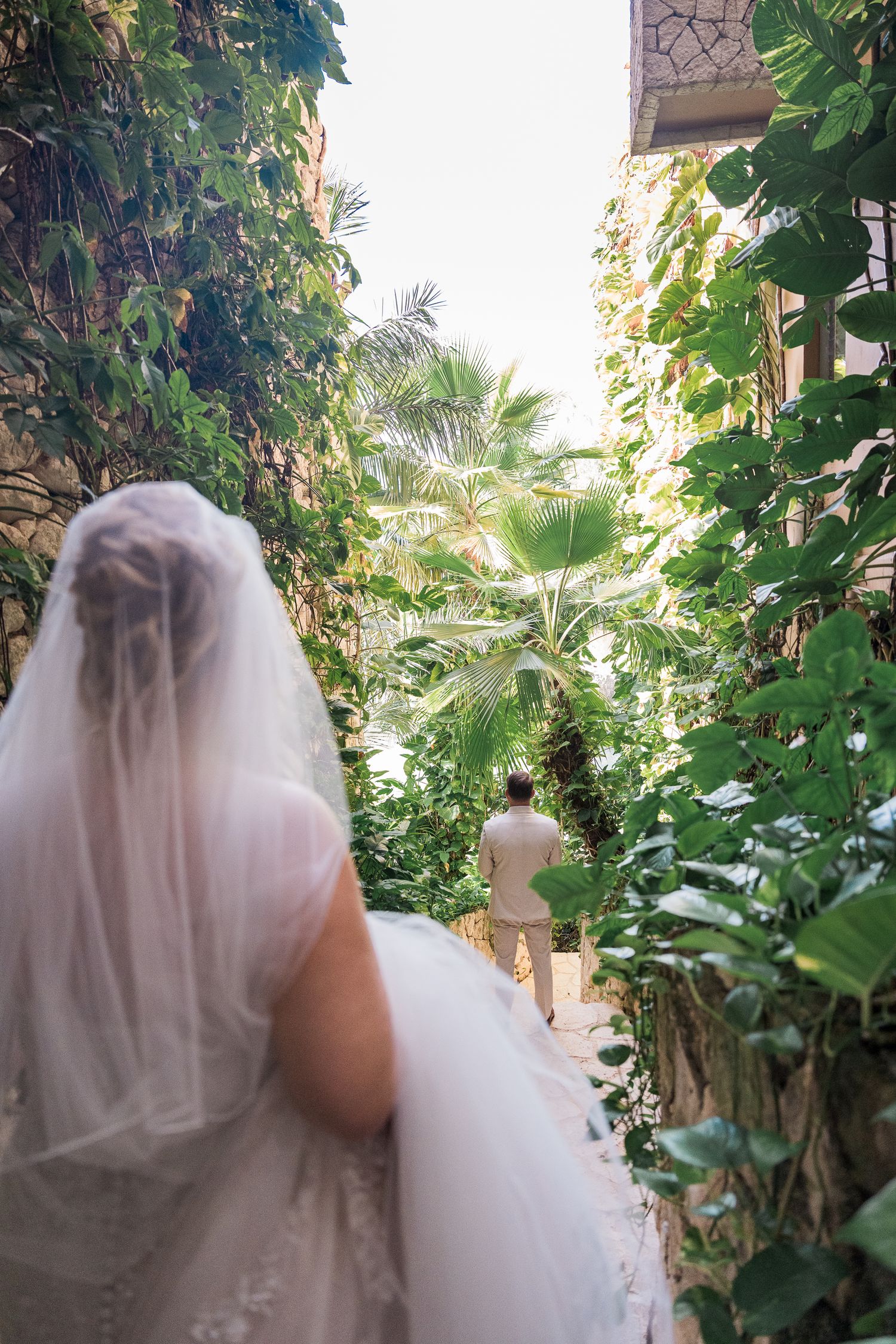 Bride in white gown and veil walks toward groom in lush tropical garden pathway on wedding day.