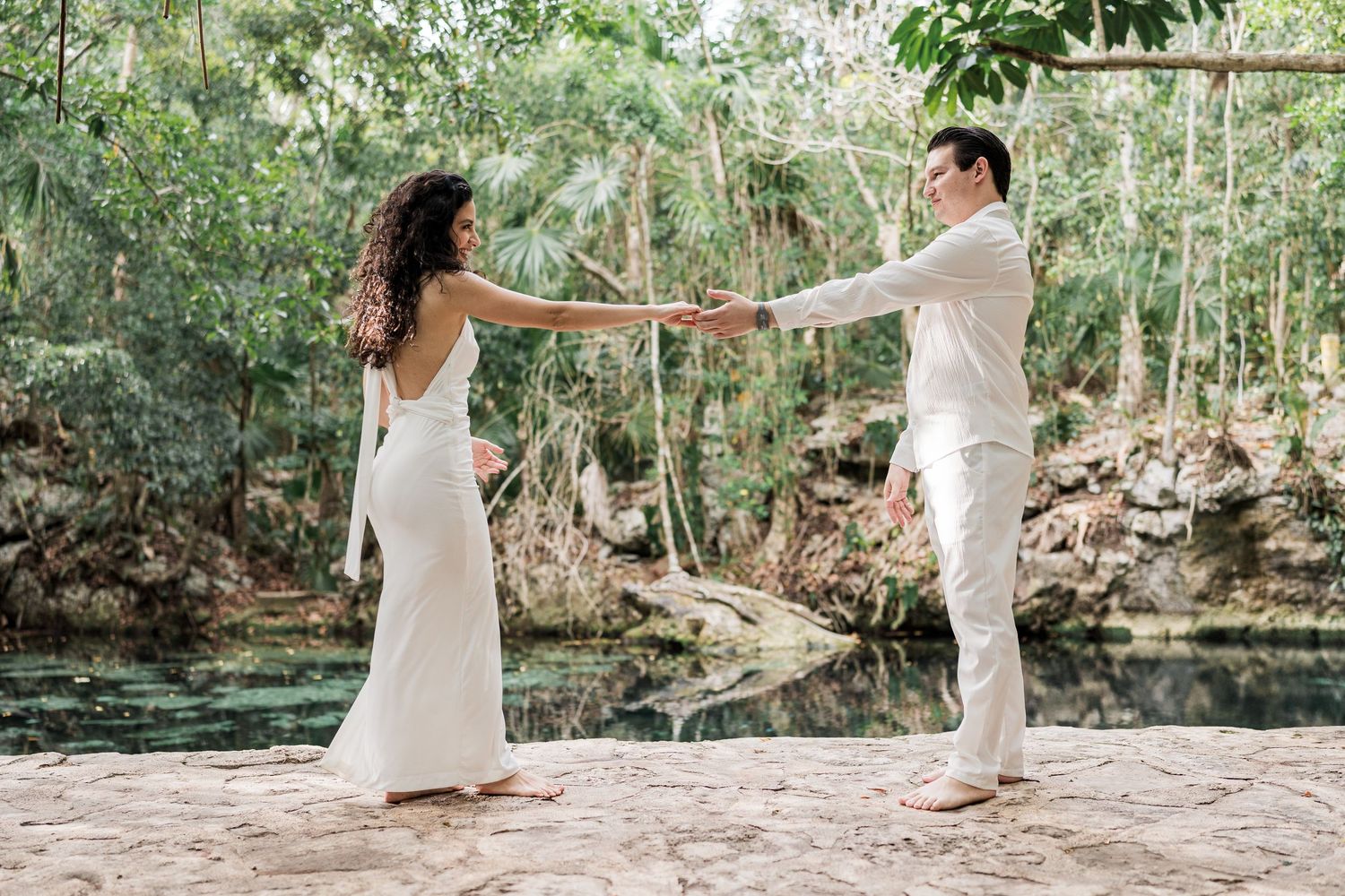Couple in white wedding attire holding hands near a lush green cenote in a tropical forest setting.