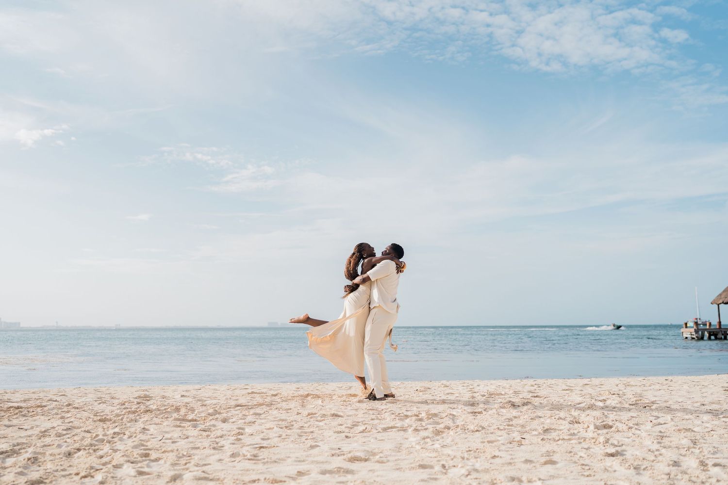 Couple embracing on a sandy beach with blue ocean and sky backdrop.