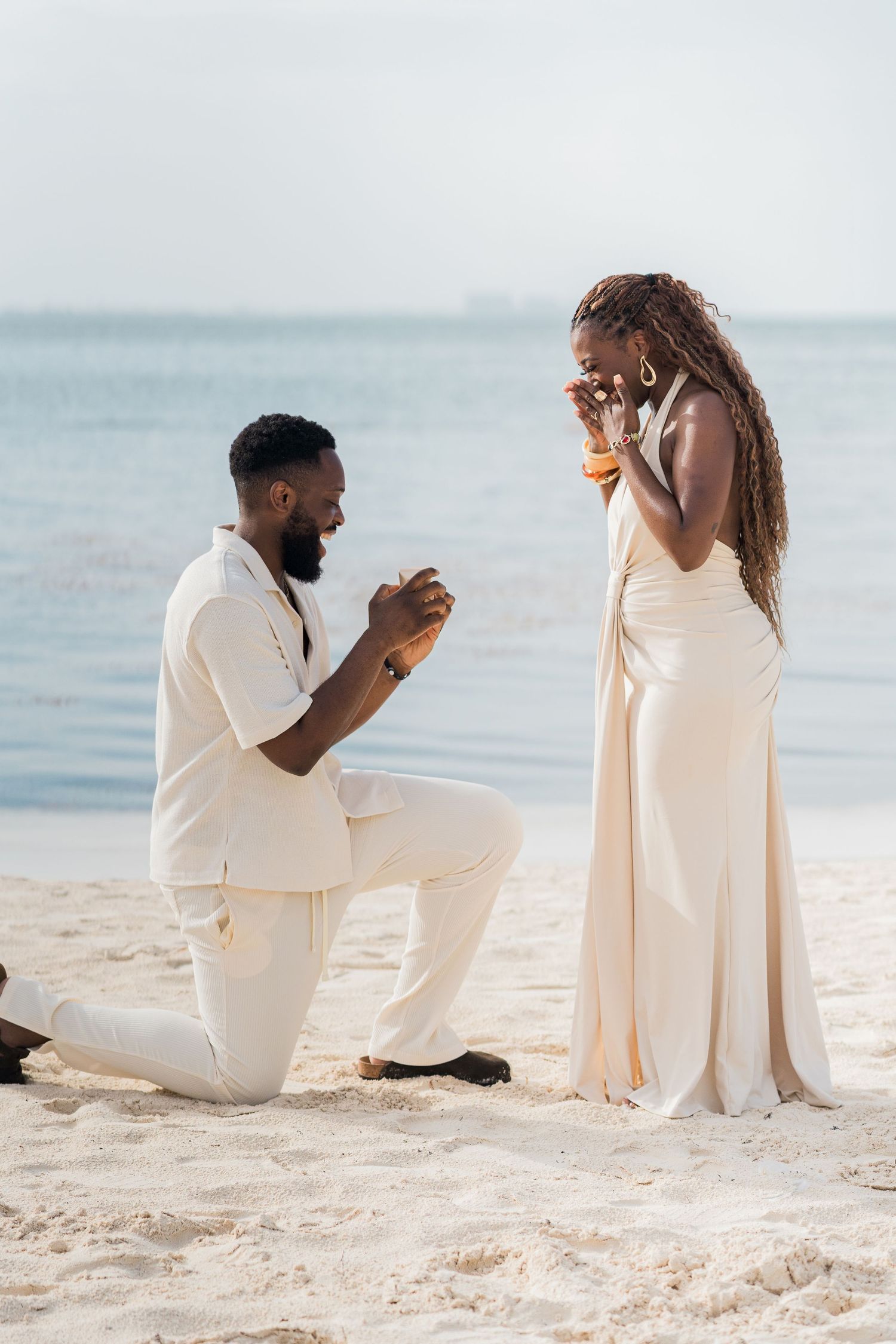 Man proposes to woman on one knee on a sandy beach, both dressed in elegant cream outfits with ocean backdrop.