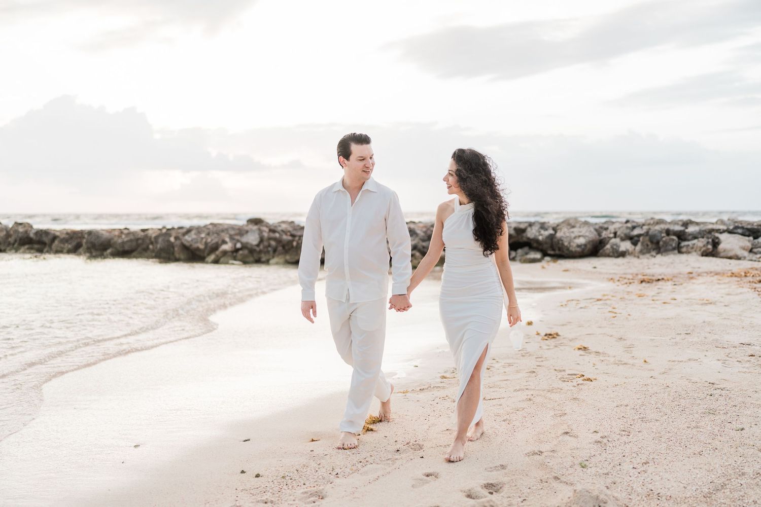 Couple holding hands walking on a sandy beach in white outfits with rocky shoreline and cloudy sky.